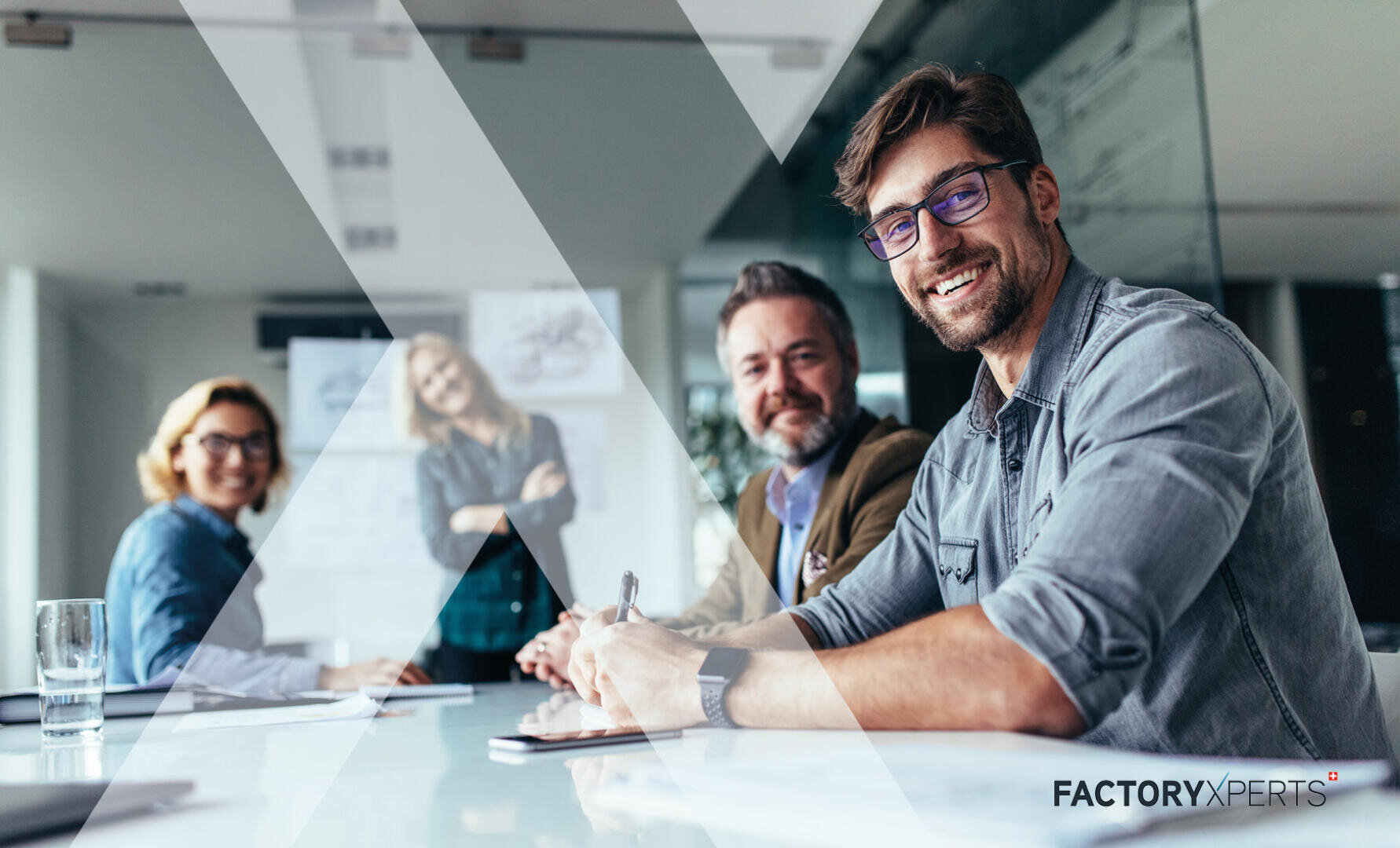 Vier lächelnde Kollegen sitzen und stehen um einen gläsernen Konferenztisch in einem modernen Büro. Ein Mann davor blickt mit einem Stift in die Kamera. Unten rechts ist das FACTORYXPERTS-Logo zu sehen.
