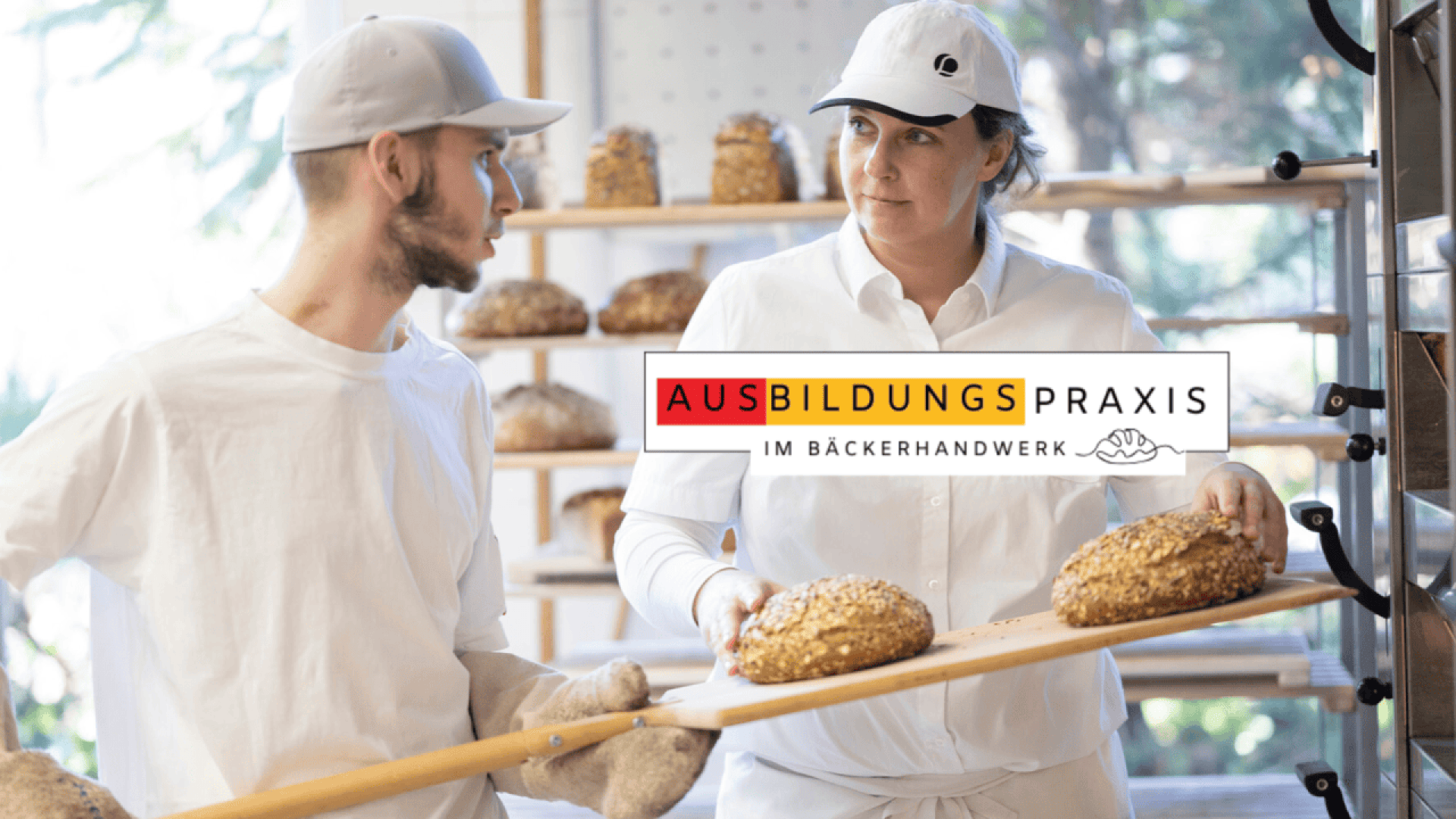 Two bakers in white uniforms hold a wooden baking peel with loaves of bread in a bakery. The logo AUSBILDUNGSPRAXIS IM BÄCKERHANDWERK is overlaid in the center of the image.