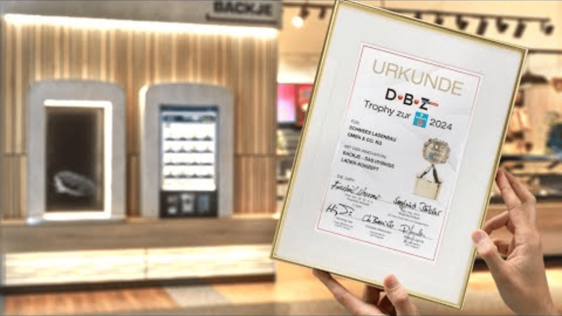Eine Person hält eine gerahmte Urkunde mit deutschem Text und Unterschriften vor einem modernen, gut beleuchteten Verkaufsautomaten und einer Verkaufsfläche.