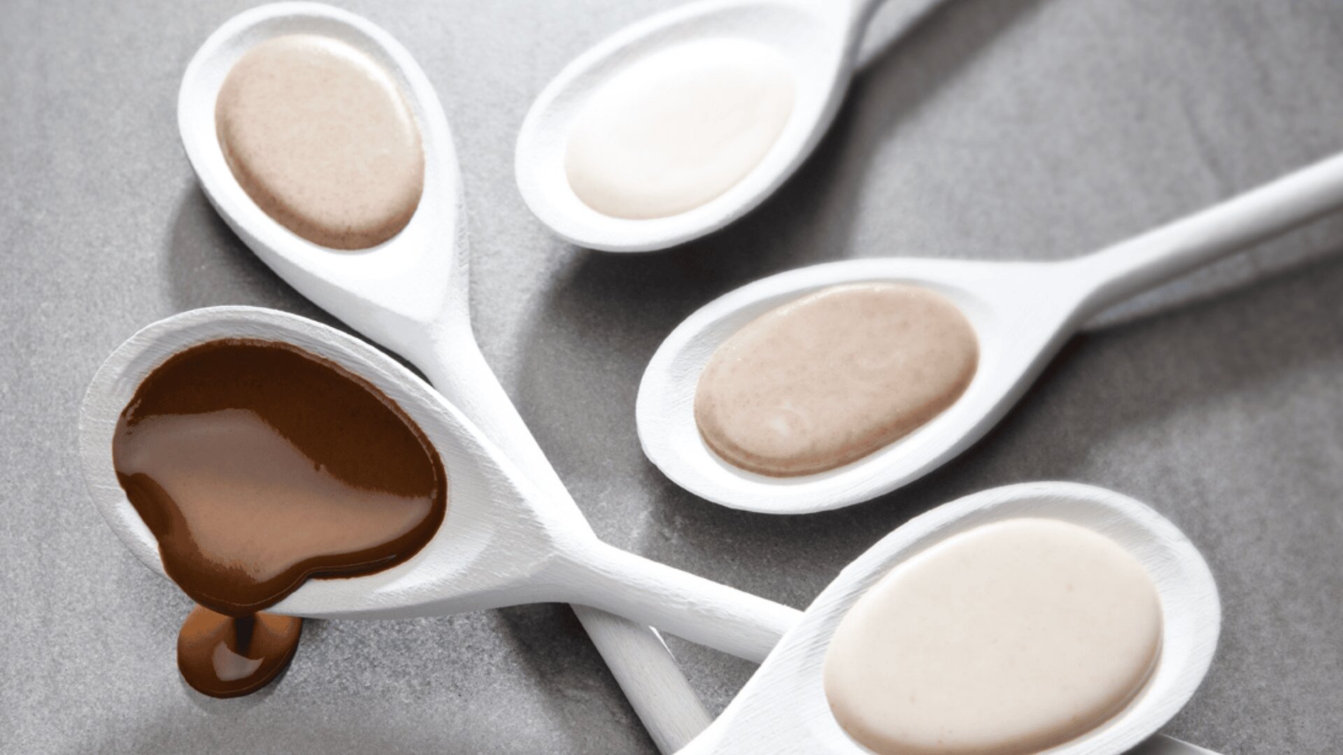Vier weiße Löffel auf einer grauen Fläche, die jeweils einen anderen cremigen Aufstrich halten. Ein Löffel hat einen braunen Aufstrich, der über den Rand tropft, während die anderen beige und cremefarbene Aufstriche enthalten.