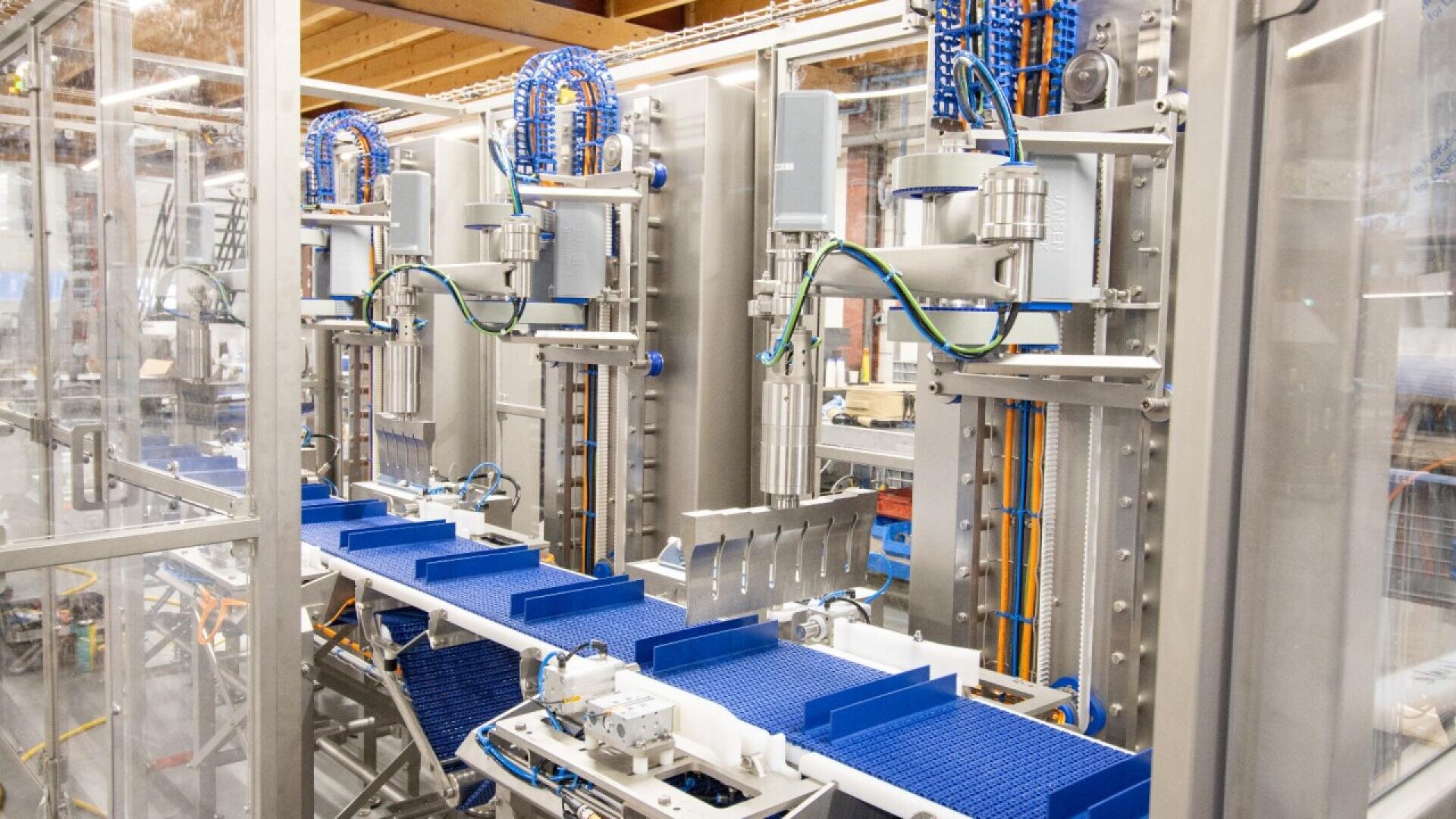 Ein modernes automatisiertes Fließband mit blauen Förderbändern, Roboterarmen und verschiedenen Maschinenkomponenten in einer gläsernen Industrieanlage.