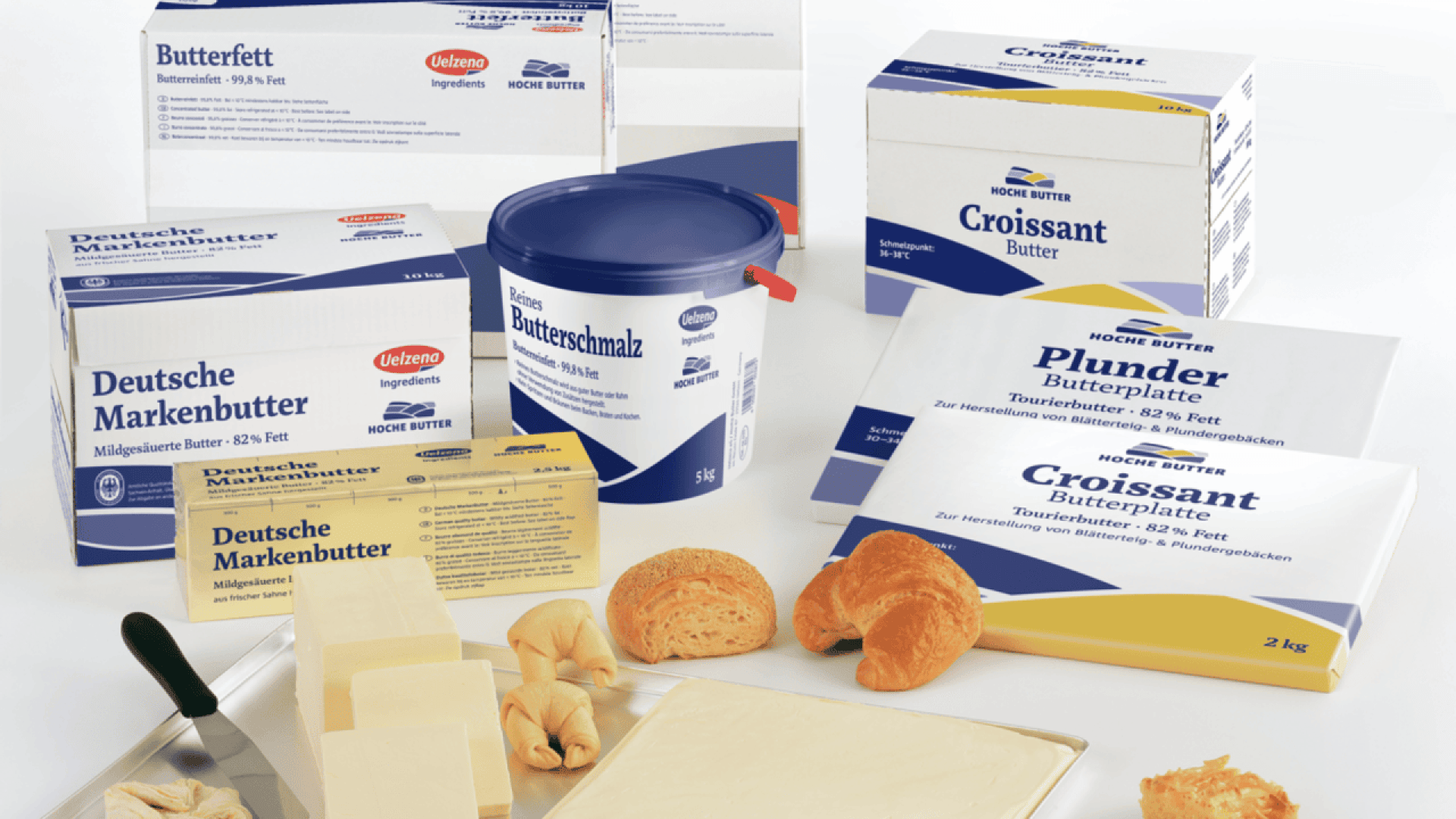 A variety of packaged German butter, margarine, and pastry products are displayed alongside unwrapped butter blocks, croissants, and a butter knife on a white surface.