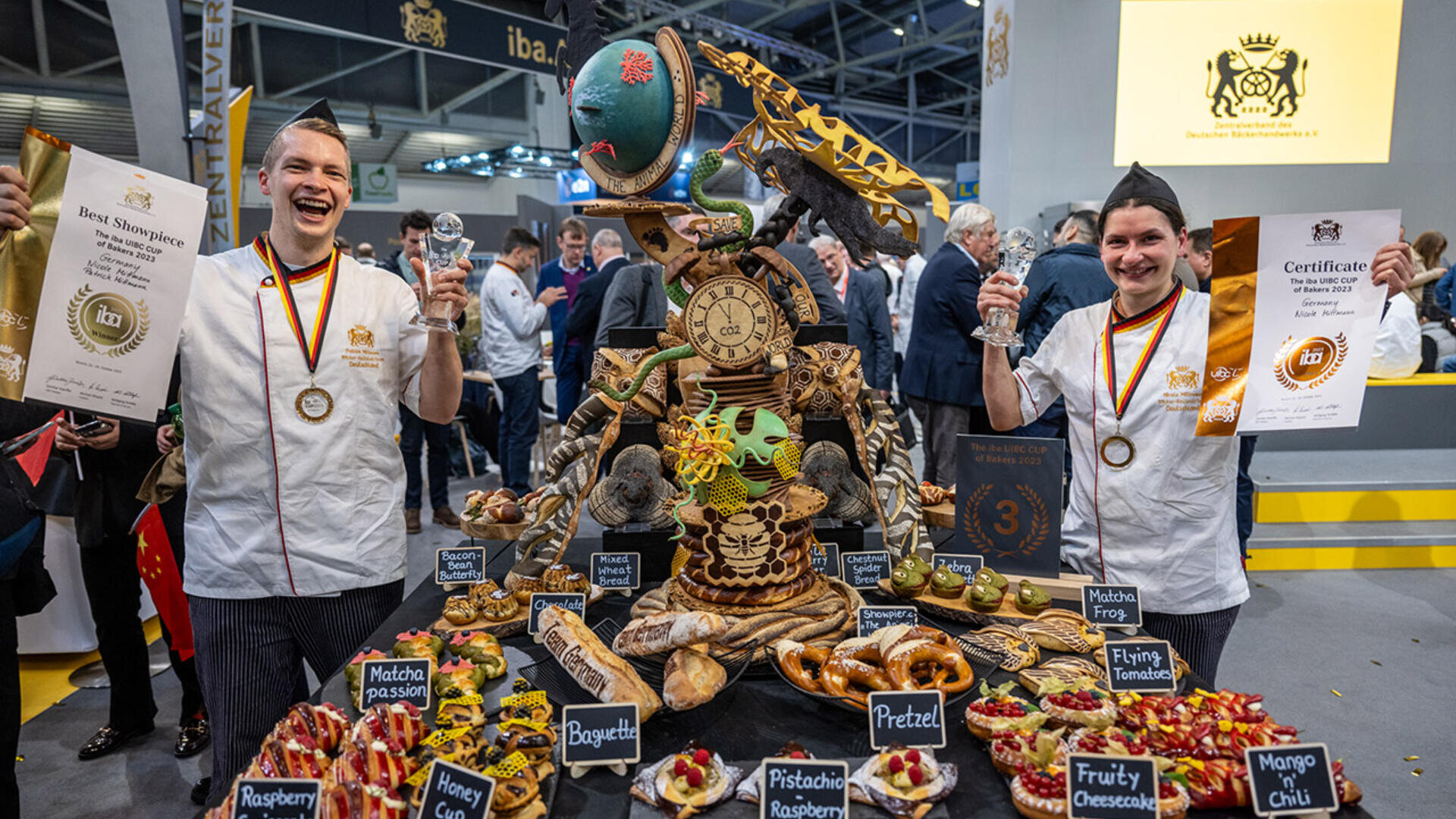 Two smiling chefs in white uniforms and medals hold trophies and certificates beside an elaborate pastry and bread display at a competition, surrounded by assorted baked goods and labeled desserts. People and banners are in the background.