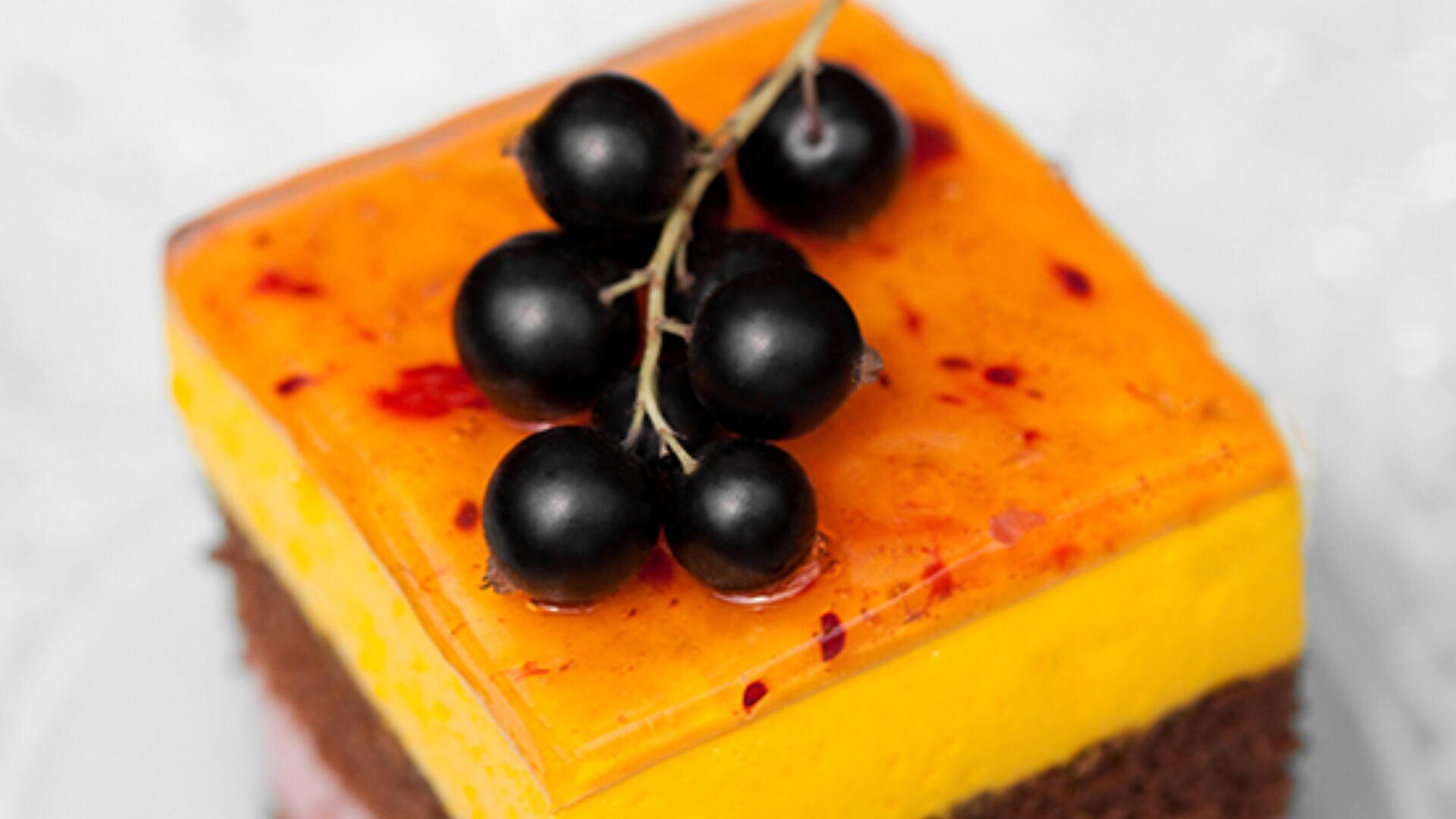 A square dessert with a yellow-orange glazed top layer, a chocolate cake base, and a cluster of blackcurrants on top, served on a white plate.