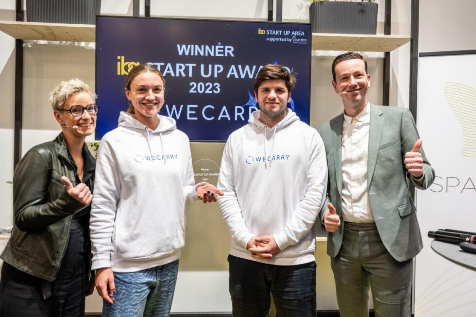 Four people stand smiling, two holding trophies, in front of a screen that reads “Winner Start Up Award 2023 WeCarry.” Three give thumbs up, and two wear white WeCarry hoodies, celebrating their achievement.
