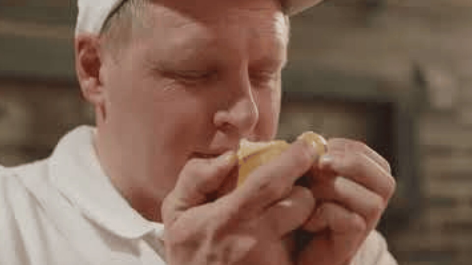 Ein Mann in weißem Hemd und Mütze hält ein kleines Stück Brot vor sein Gesicht und riecht zufrieden daran. Der Hintergrund ist unscharf.