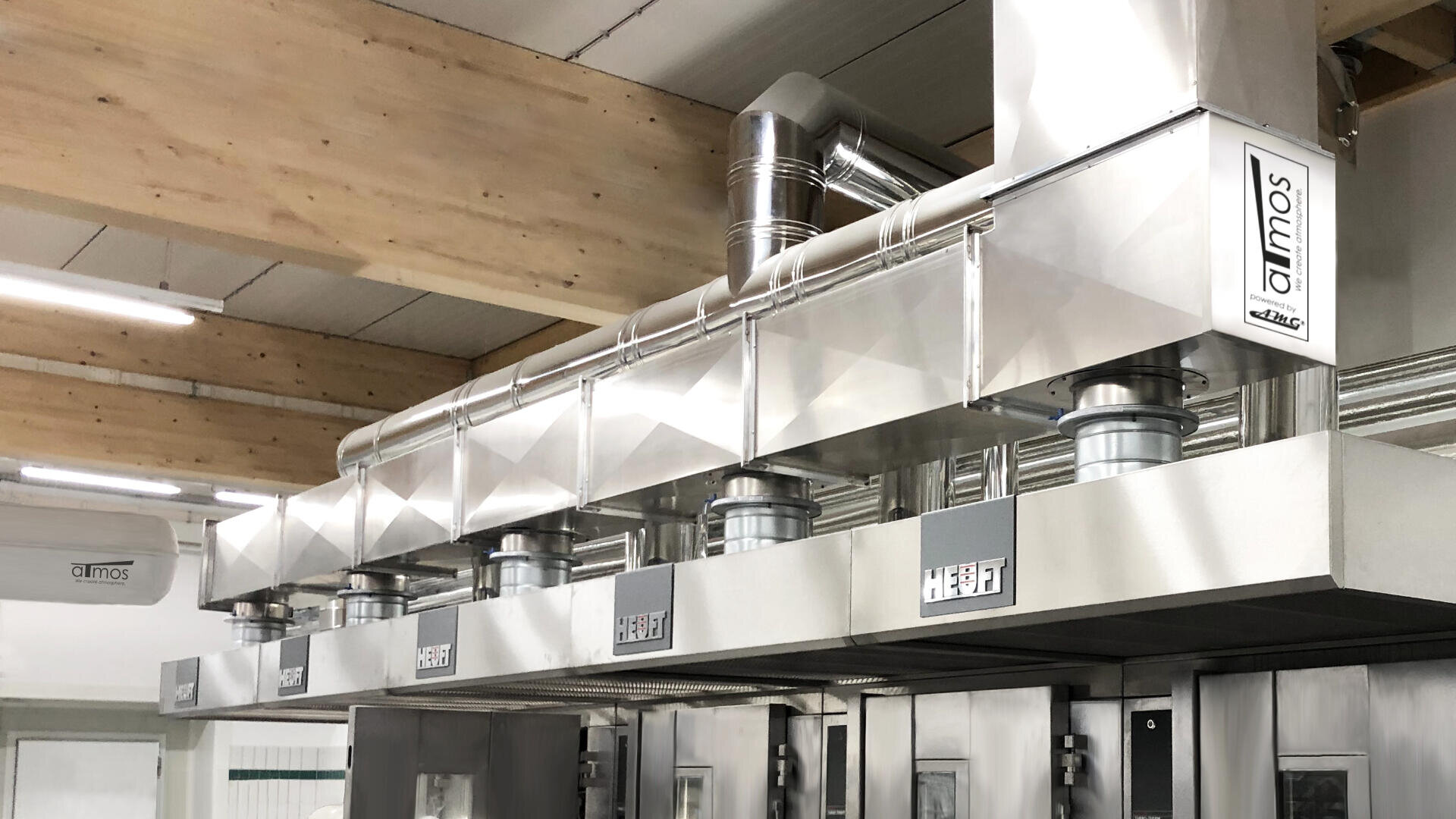 Eine Reihe von industriellen Edelstahl-Lüftungshauben mit Kanälen, die über gewerblichen Öfen in einer modernen Lebensmittelproduktionsanlage installiert sind.