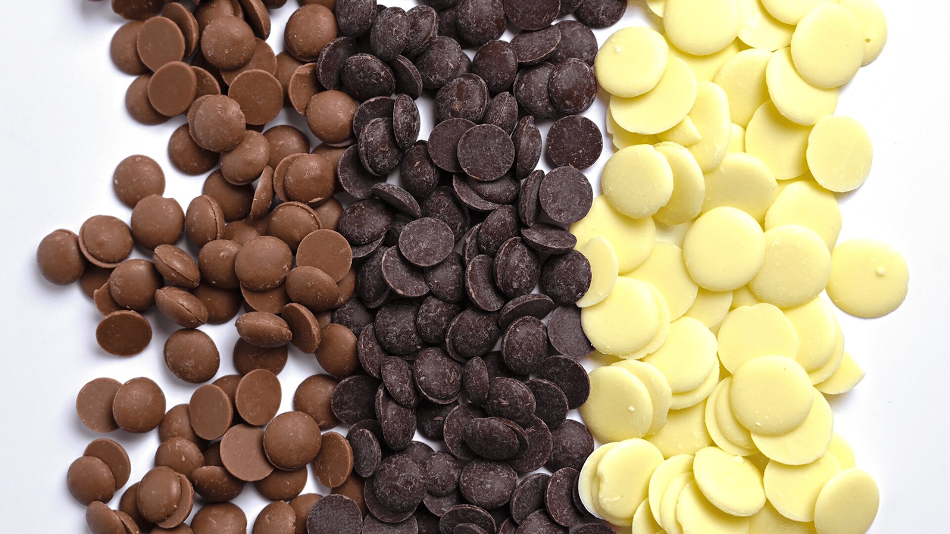 Three piles of chocolate chips—milk chocolate, dark chocolate, and white chocolate—are arranged side by side on a white surface.