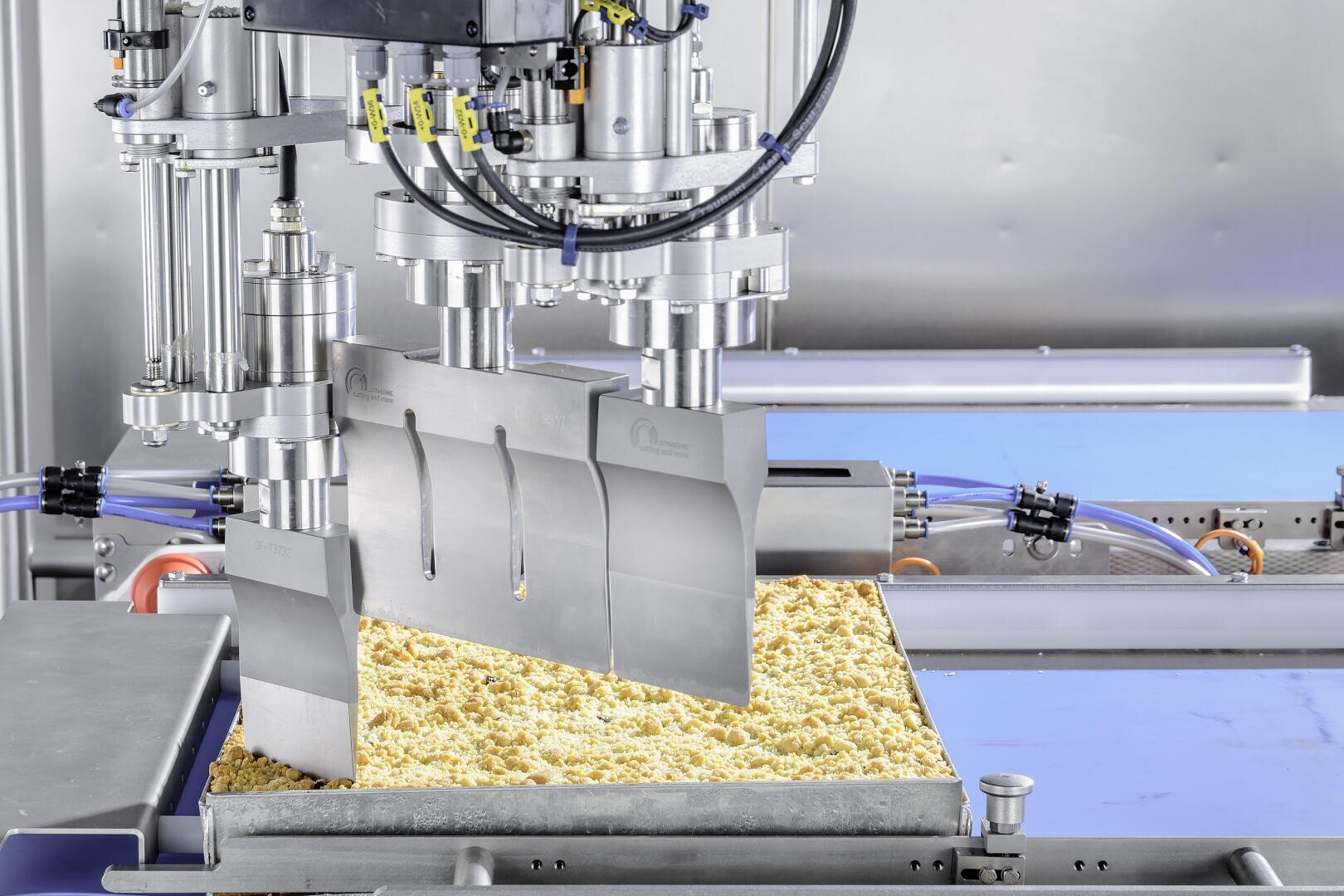 Eine Nahaufnahme einer Lebensmittelverarbeitungsmaschine, die in einer industriellen Umgebung ein großes Tablett mit krümeligem, gelbem Teig oder Lebensmittel in Stücke schneidet.