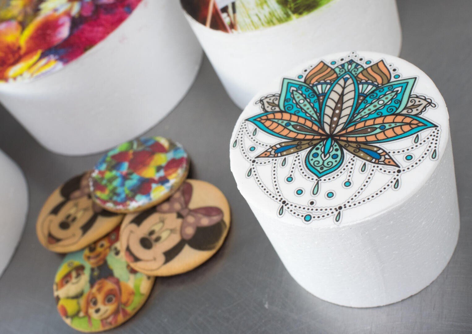 Round cakes and cookies with colorful edible printed designs, including a detailed blue and orange mandala, cartoon characters, and abstract patterns, displayed on a gray surface.