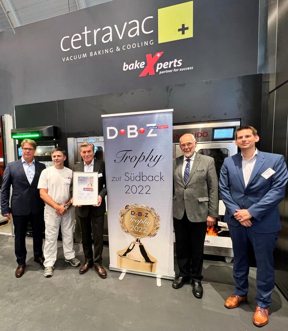 Fünf Männer in Geschäfts- und Arbeitskleidung stehen neben einem Banner mit der Aufschrift DBZ Trophy zur Südback 2022 vor einer CETRAVAC Back- und Kühlkabine. Ein Mann hält eine gerahmte Urkunde in der Hand.