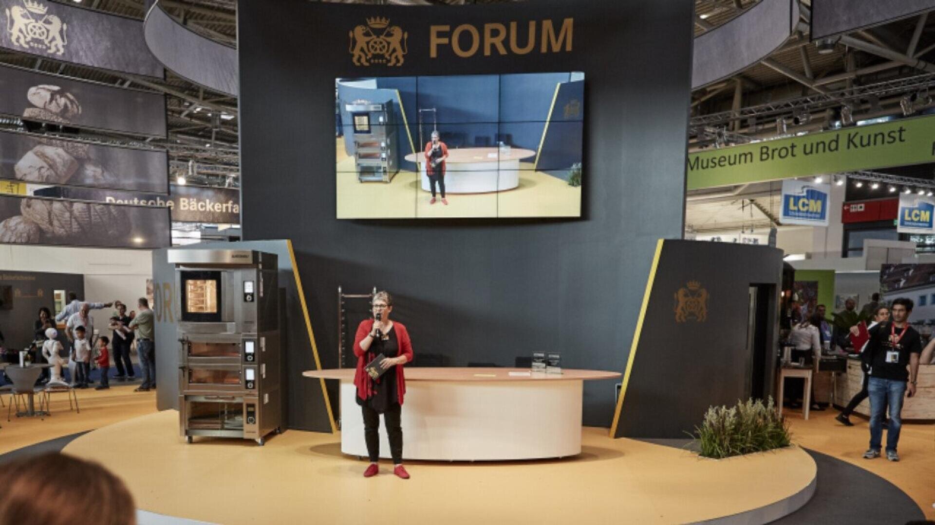 A woman stands and speaks with a microphone on a stage labeled “FORUM” at a trade show, with a large screen behind her displaying her image. There are booths, signs, and attendees in the background.
