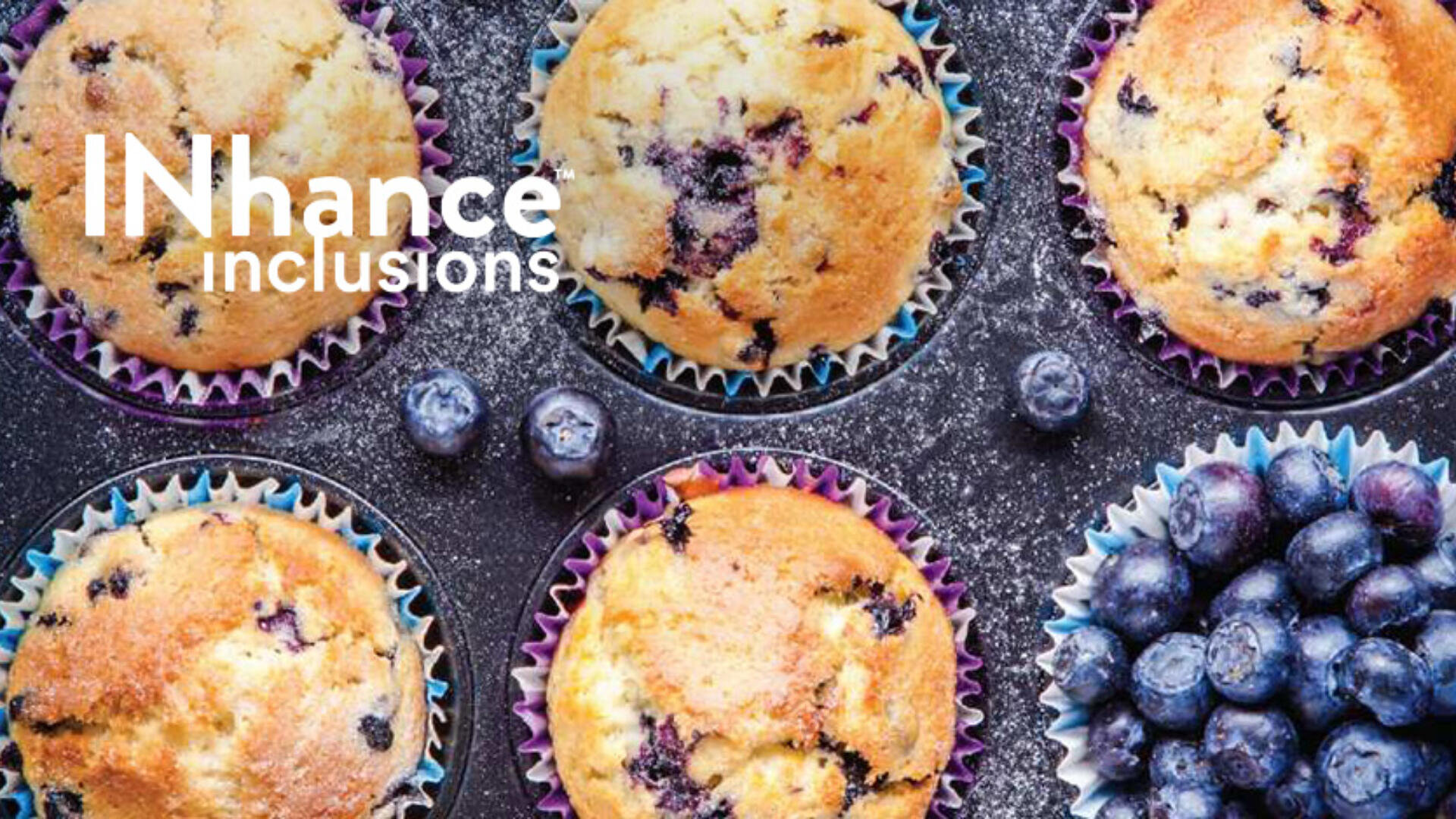 Overhead view of blueberry muffins in colorful wrappers with fresh blueberries scattered around. Text reads “INhance inclusions” in white letters on the upper left side.