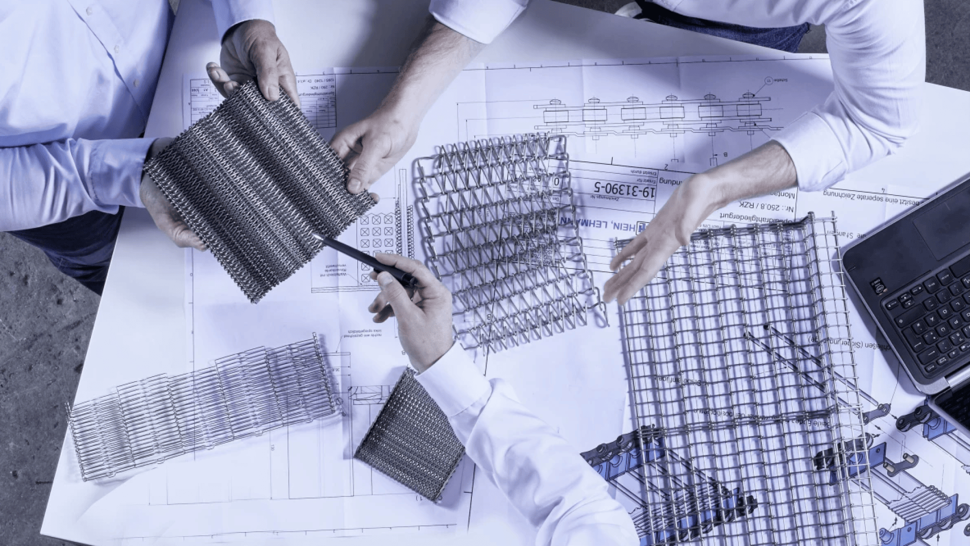 Drei Personen halten und untersuchen Muster von Metallgeweben über technischen Plänen auf einem Schreibtisch, auf dem auch ein Stift, ein Laptop und weitere Gewebestücke zu sehen sind.