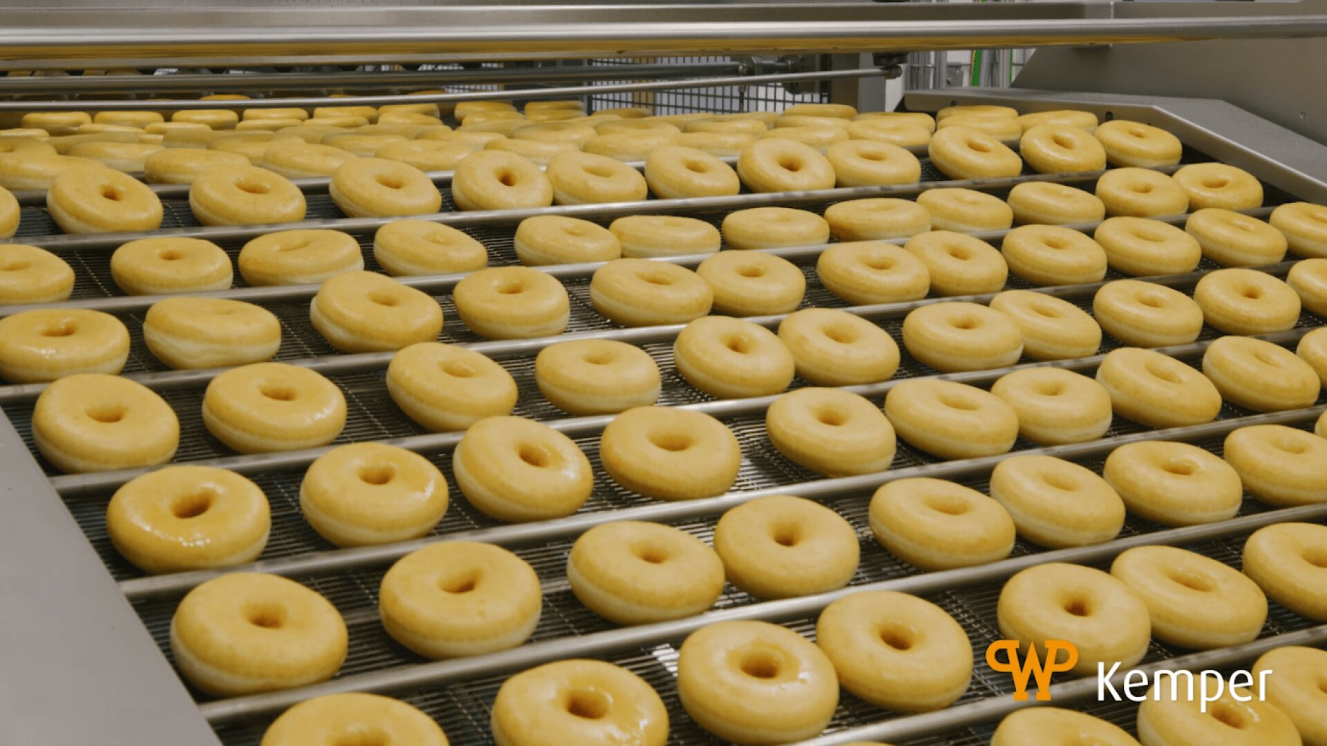 Rows of raw doughnuts are lined up on a metal conveyor belt in a factory setting, ready for baking or frying. The Kemper company logo appears in the lower right corner.