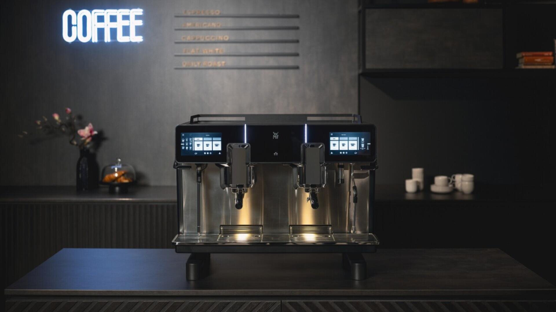 A modern, double-spout coffee machine with digital displays sits on a countertop in a sleek, dark kitchen. A neon COFFEE sign glows on the wall, and cups and decor are visible in the background.