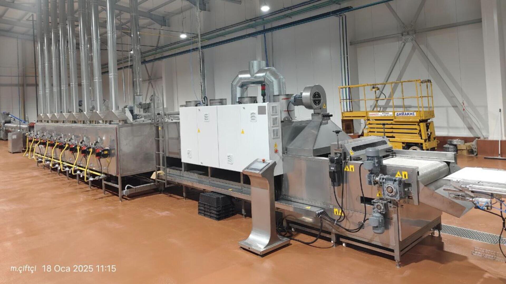 Eine große industrielle Lebensmittelverarbeitungsmaschine mit Metallkomponenten und Bedienfeldern steht in einer sauberen Fabrik mit braunem Boden und hoher Decke. Im Hintergrund ist ein gelber Wartungsaufzug zu sehen.