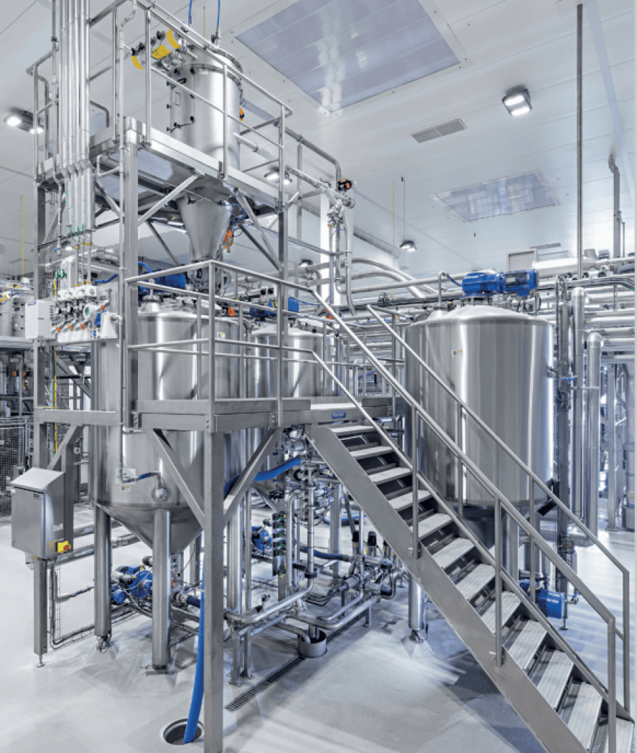 Eine moderne Industrieanlage mit großen Edelstahltanks, Rohren und Metalltreppen, die wahrscheinlich für die Verarbeitung von Lebensmitteln oder Getränken genutzt wird, mit heller Beleuchtung und sauberer, ordentlicher Umgebung.