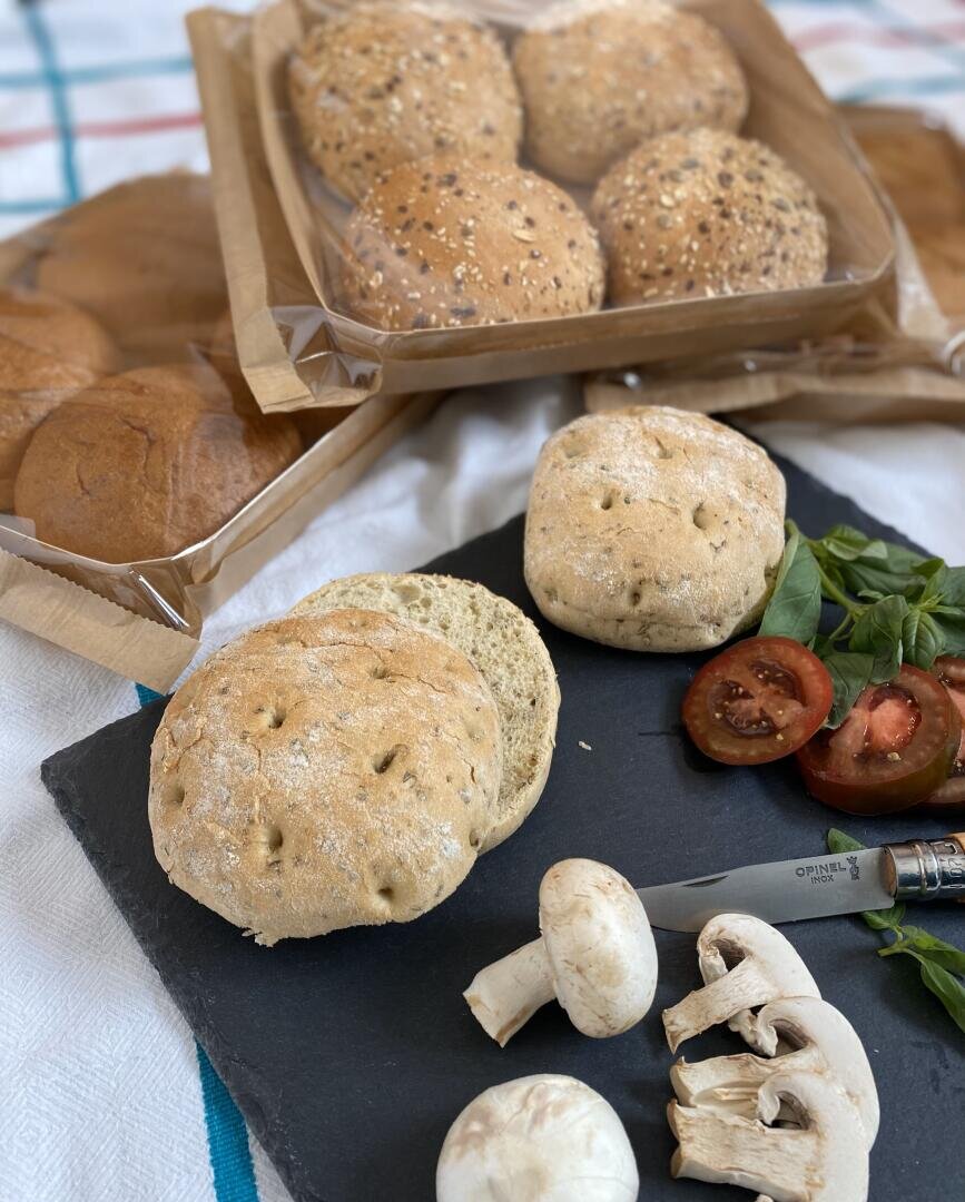 A slate board with two seeded bread rolls, sliced mushrooms, tomato slices, greens, and a knife. In the background, two trays hold more seeded and plain bread rolls on a checkered cloth.