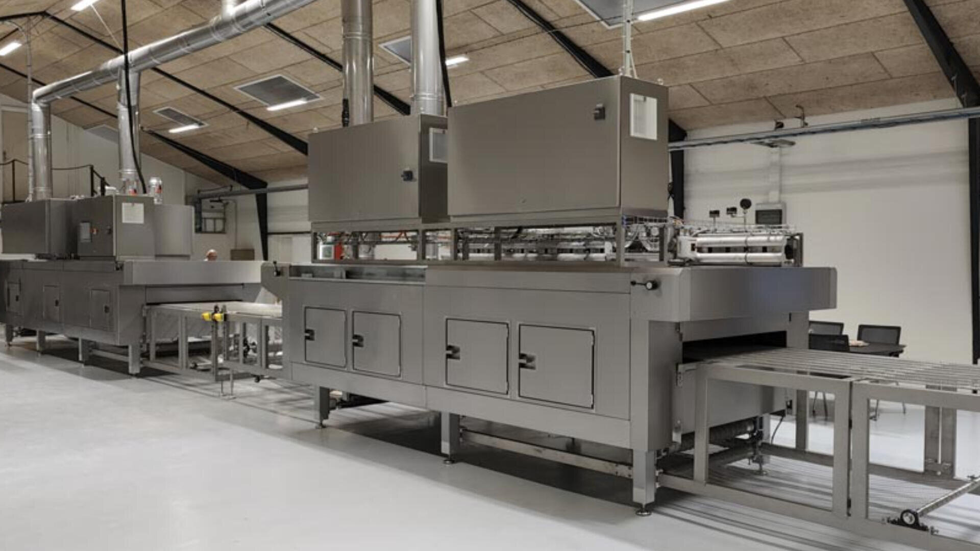 Large industrial machine with metal conveyor belts and ventilation ducts, situated in a spacious, modern factory setting with white walls and a tiled ceiling.