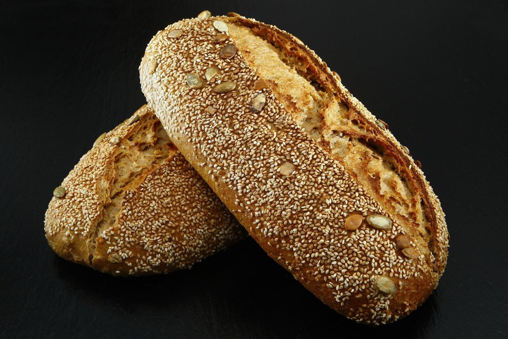 Two rustic loaves of bread topped with sesame and sunflower seeds rest on a dark surface, with one loaf leaning diagonally on the other.