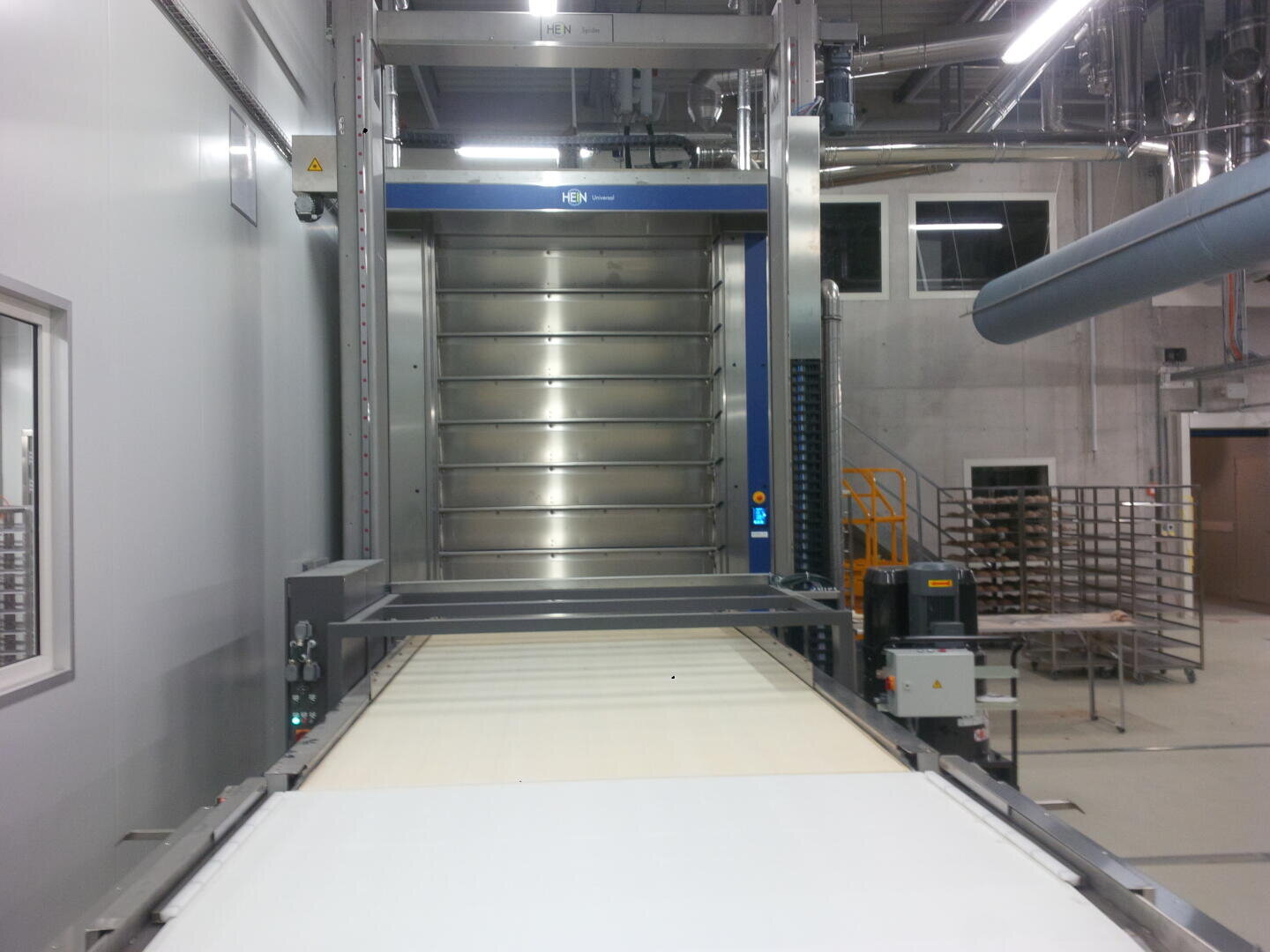 Ein großer industrieller Backofen mit mehreren Regalen steht am Ende eines Förderbands in einer sauberen, modernen Fabrikumgebung mit Metallgestellen und Geräten im Hintergrund.