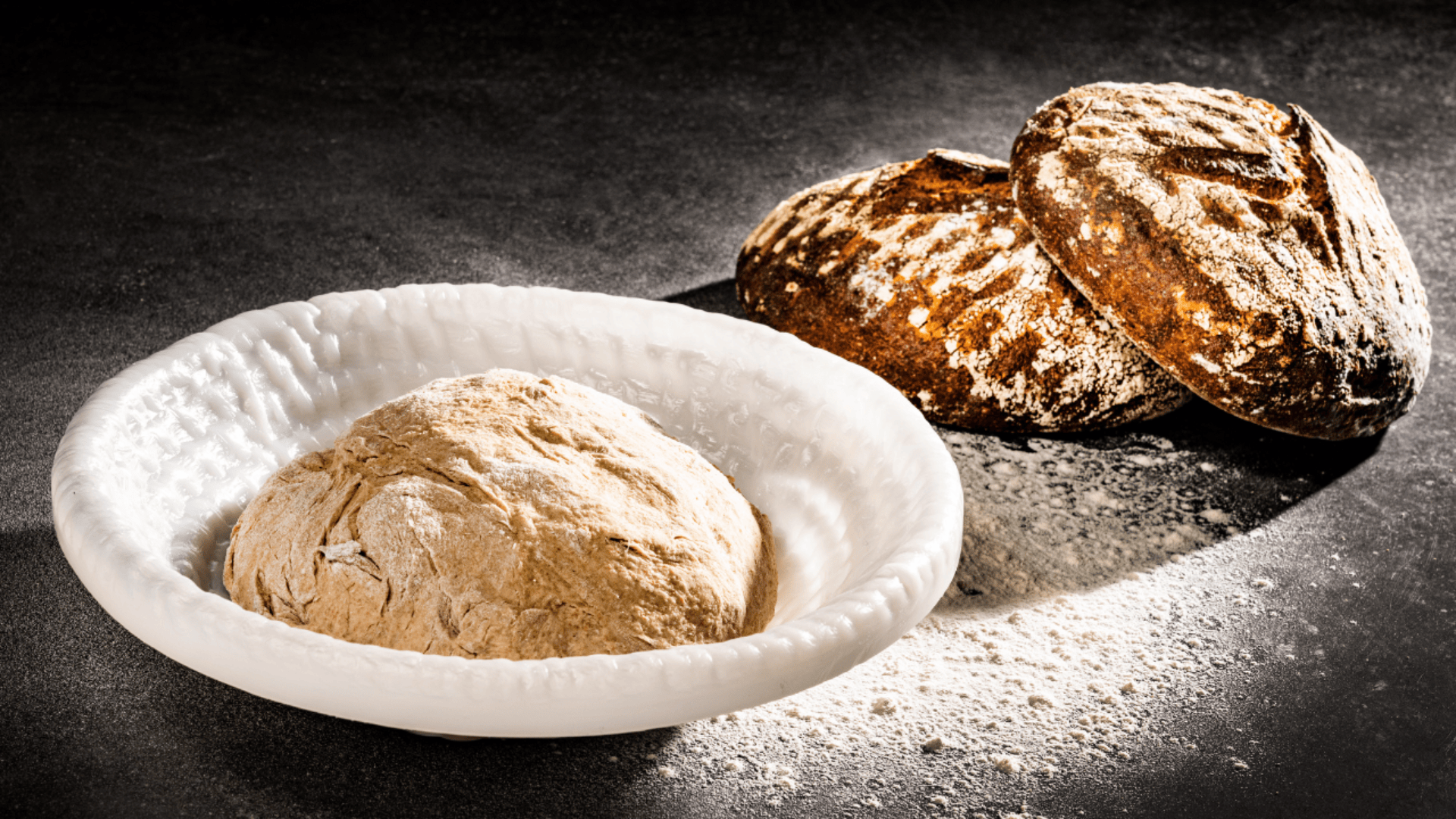 Eine Kugel aus rohem Brotteig in einer weißen Schüssel liegt auf einer dunklen, mit Mehl bestäubten Fläche, während im Hintergrund zwei runde, rustikale Brote gebacken werden.