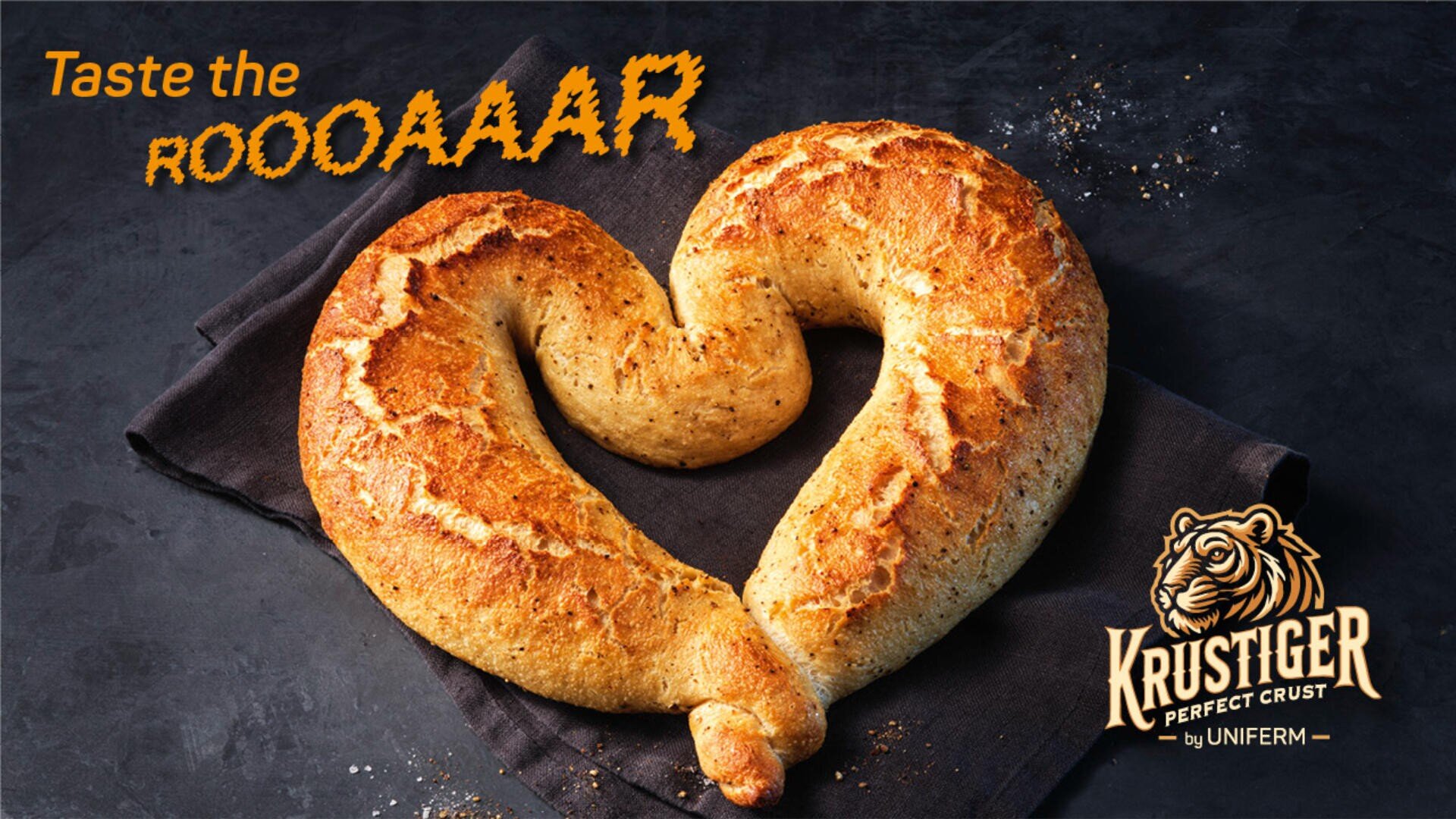 Ein herzförmiger, goldbrauner Laib Brot liegt auf einem dunklen Tuch. Der Text lautet: „Taste the ROOOAAAR“. Unten rechts ist ein Tiger-Logo mit Krustiger Perfect Crust von Uniferm zu sehen.