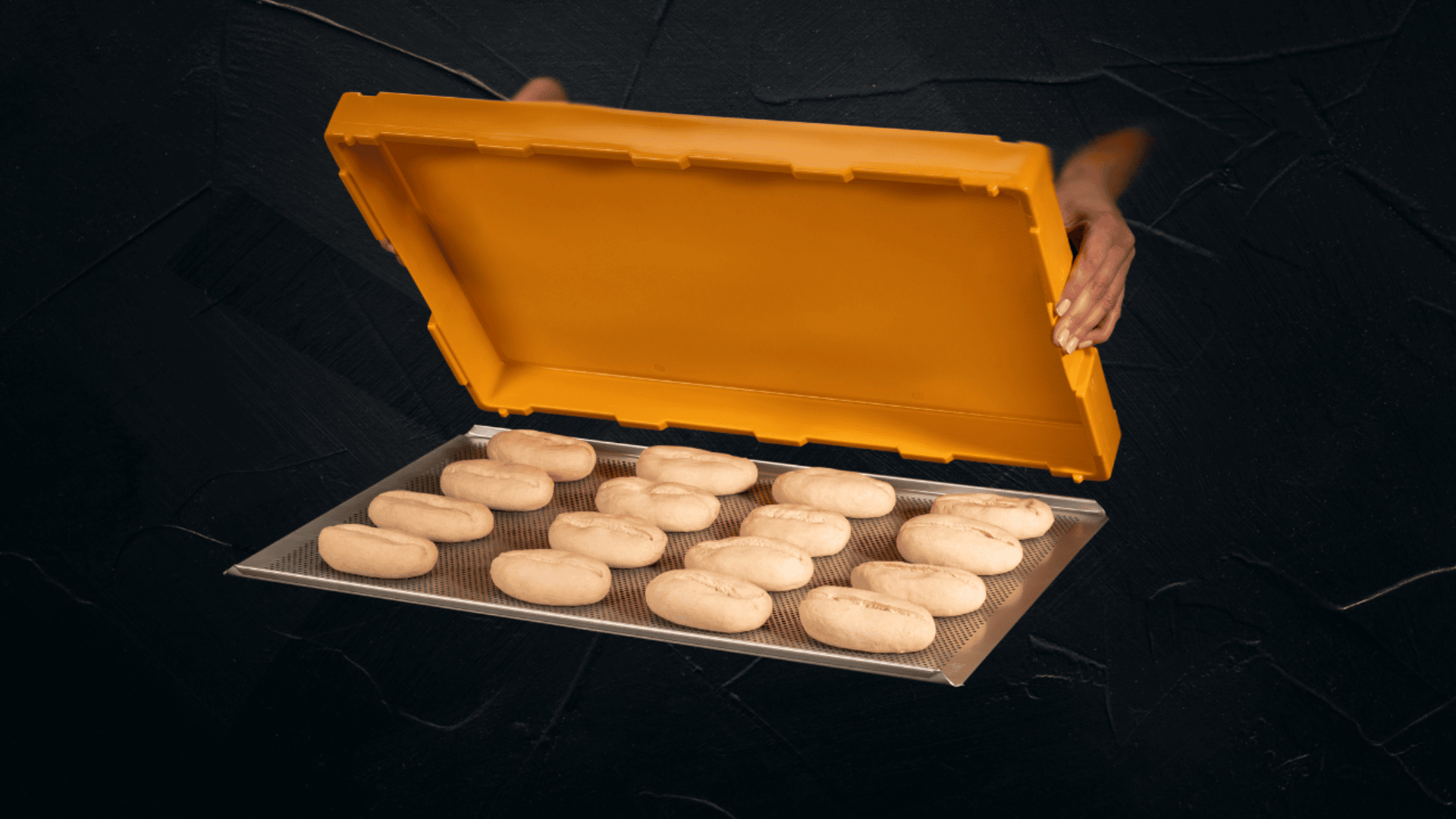 Die Hände heben den orangefarbenen Deckel von einem Tablett mit Reihen von ungebackenen ovalen Teiglingen, die sich vor einem dunklen Hintergrund befinden.