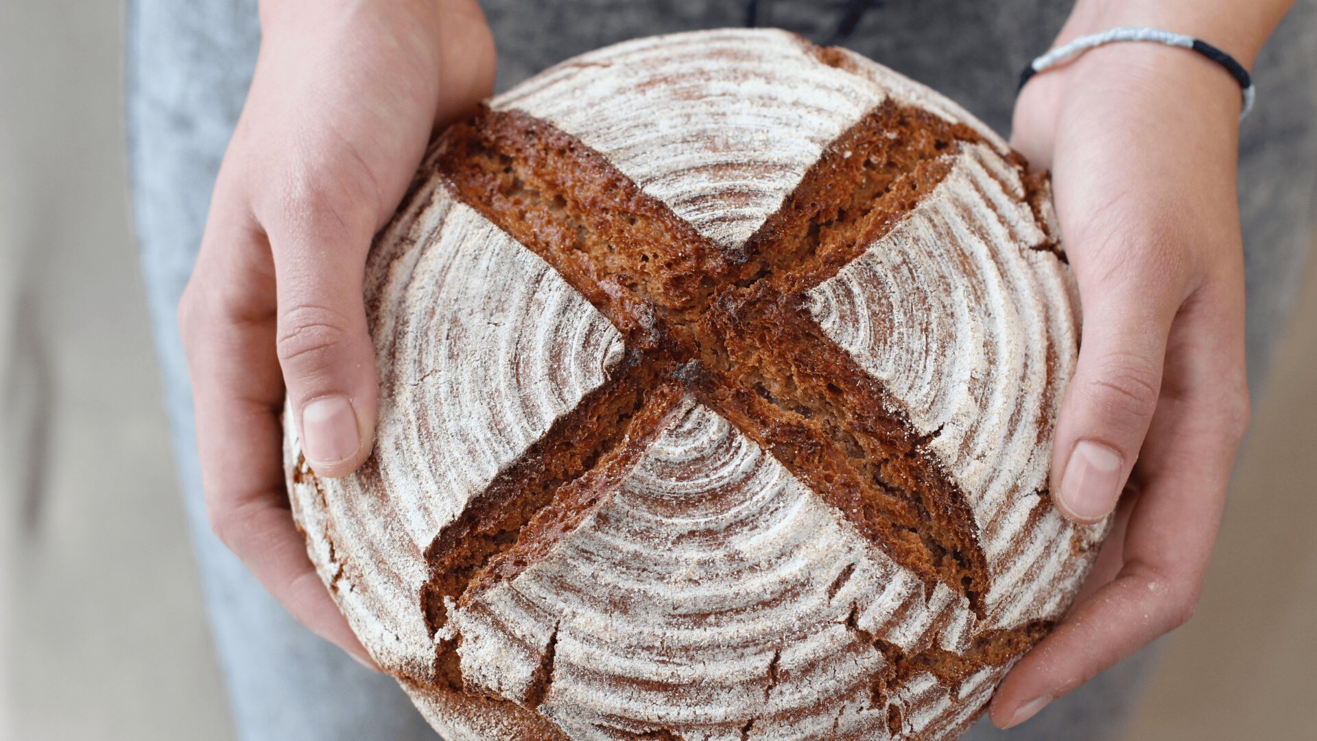 A person holding a round, rustic loaf of bread with a deep X-shaped slash on top and a dusting of flour, showing a spiral pattern from the proofing basket.