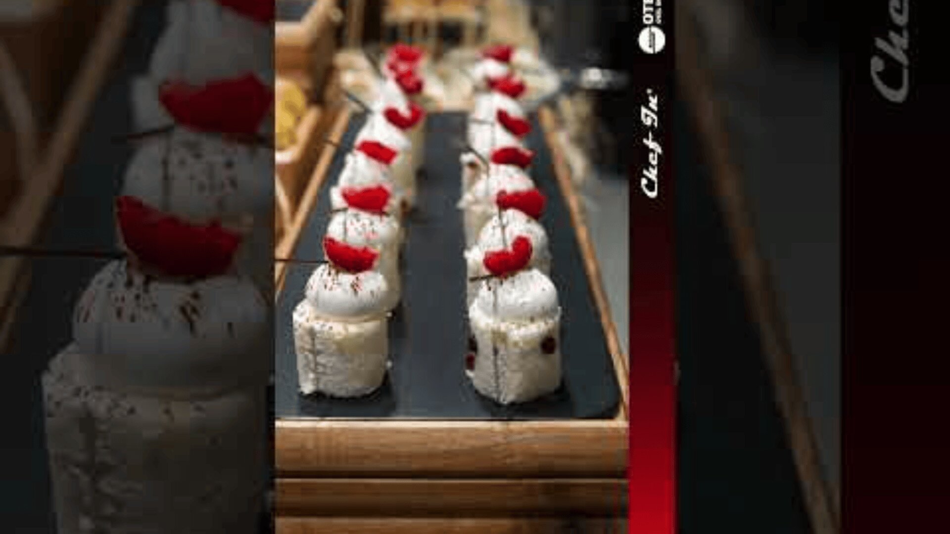 Rows of elegant white desserts topped with whipped cream and red fruit slices are arranged neatly on a black tray, displayed in a bakery or restaurant setting.