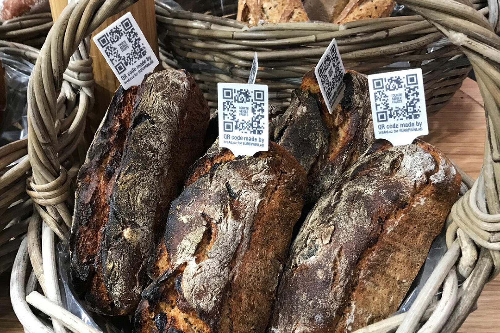 Ein Weidenkorb, gefüllt mit rustikalen Brotlaiben, die jeweils mit einem QR-Code auf einer kleinen weißen Karte beschriftet sind. Im Hintergrund sind weitere Körbe mit Brot zu sehen.