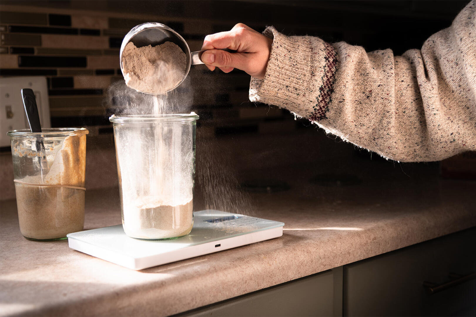 A person in a sweater pours flour from a measuring cup into a glass jar on a kitchen scale. Another jar with a spatula and brown dough is on the counter. Sunlight shines on the scene.