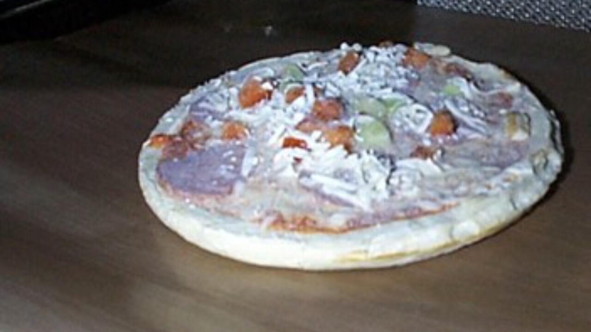 A slightly blurry, partially cooked frozen pizza topped with cheese and a few pieces of vegetables and meat sits on a brown oven tray.