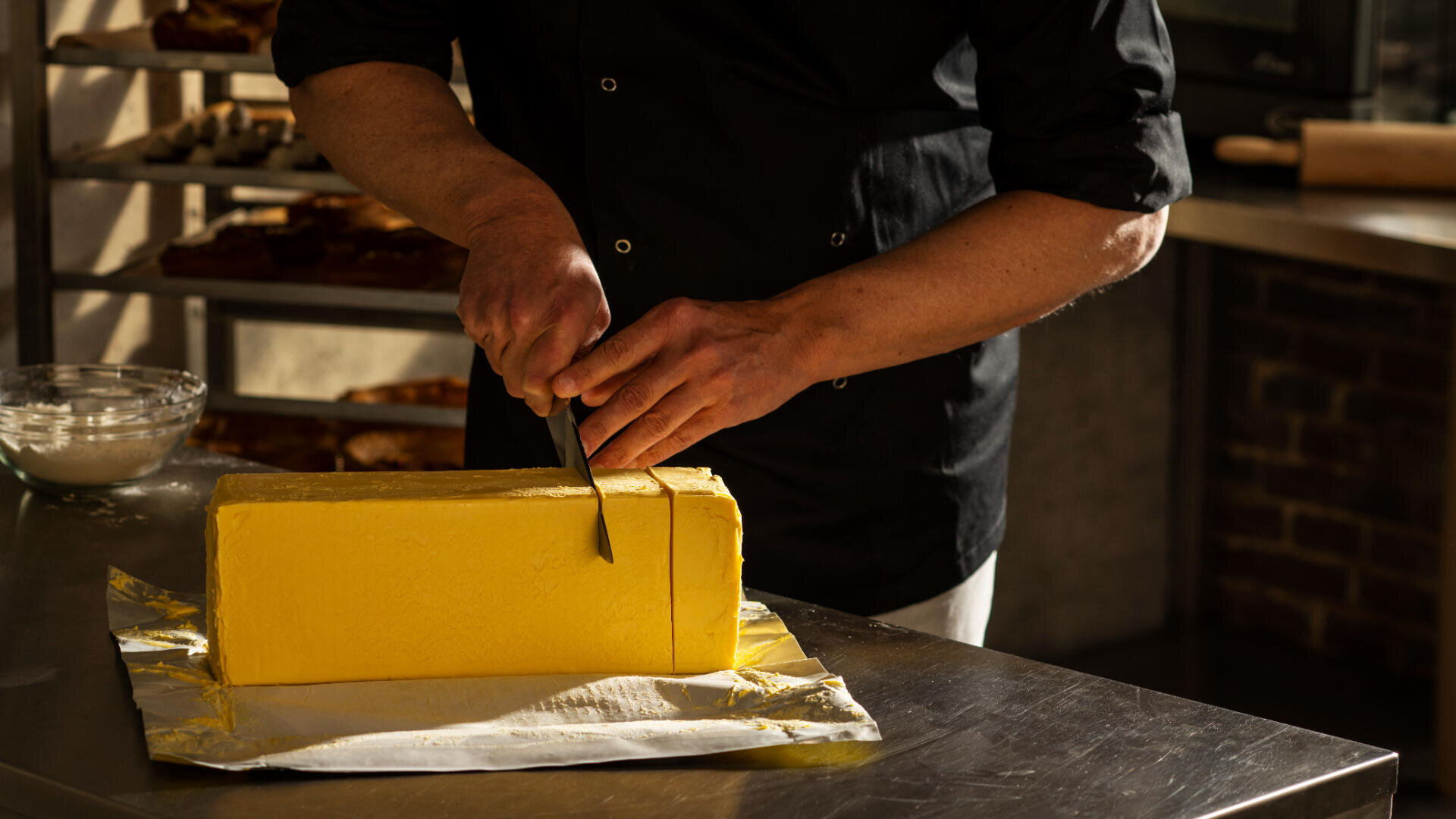 A person in a dark chef’s jacket cuts a large rectangular block of butter with a knife on a stainless steel countertop in a bakery kitchen.