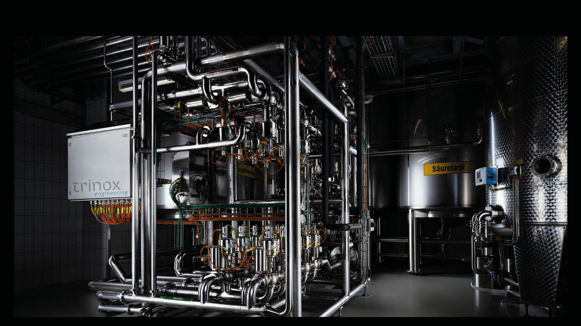 Industriemaschinen aus rostfreiem Stahl mit zahlreichen Rohren, Ventilen und Drähten, untergebracht in einer sauberen, modernen Anlage. Etiketten wie trinox engineering und Säueretank sind auf den Geräten sichtbar. Die Beleuchtung hebt die metallischen Oberflächen hervor.