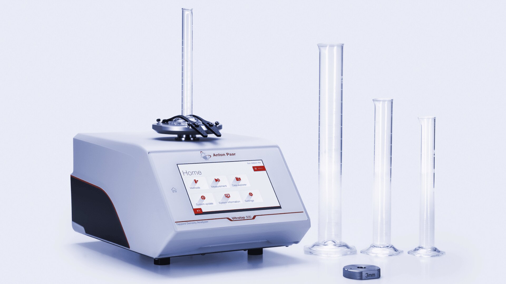 A laboratory instrument with a touchscreen display is shown next to three transparent graduated cylinders of different sizes and a small round object marked “3cm³,” all on a white surface.