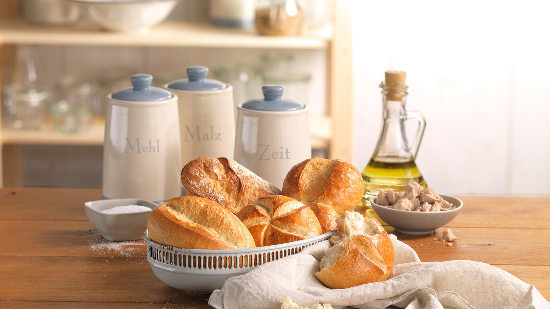 Auf einem Holztisch mit weißem Tuch steht eine Schüssel mit frisch gebackenen Brötchen. Daneben stehen Keramiken mit den Aufschriften „Mehl“, „Malz“ und „Zeit“, eine Flasche Olivenöl, ein Schälchen mit Salz und eine kleine Schüssel Hefe.