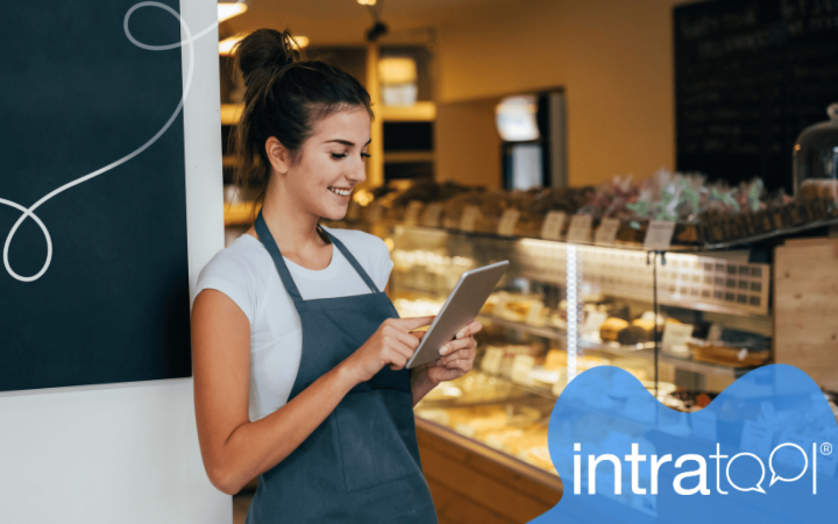 A smiling woman wearing an apron uses a tablet while standing in a bakery. Shelves with various pastries are visible in the background. The lower right corner shows a blue shape with the intratool logo.