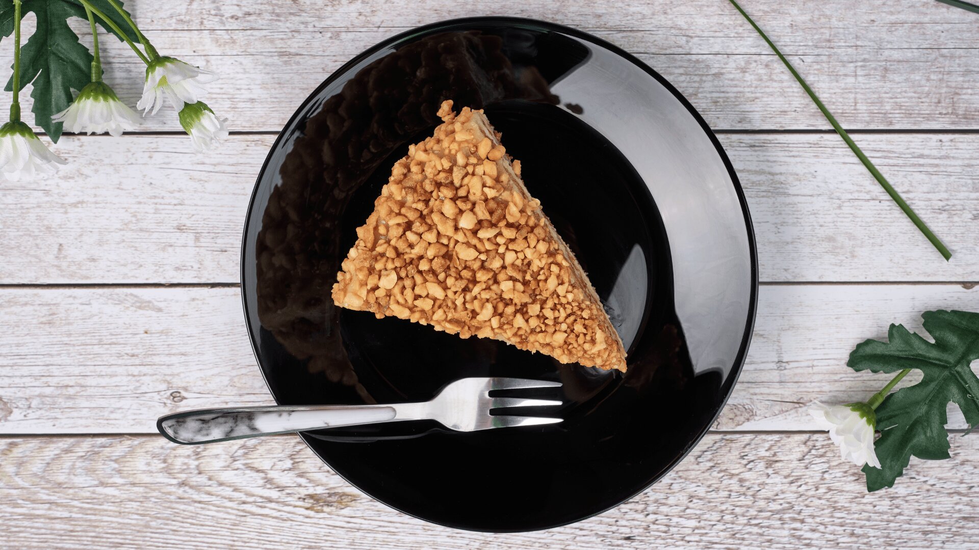 A slice of cake topped with chopped nuts is on a black plate with a fork, placed on a rustic white wooden surface. White flowers and green leaves surround the plate.