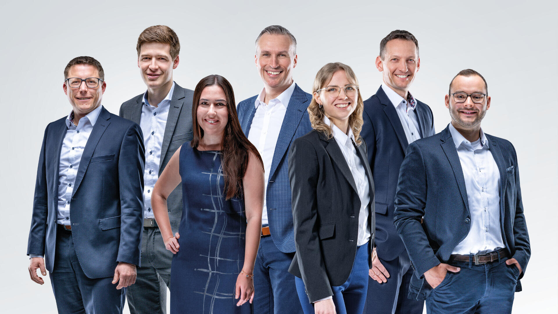 Seven people in business attire stand side by side, smiling at the camera, against a light gray background. Four men and three women are included, dressed in suits and formal outfits.