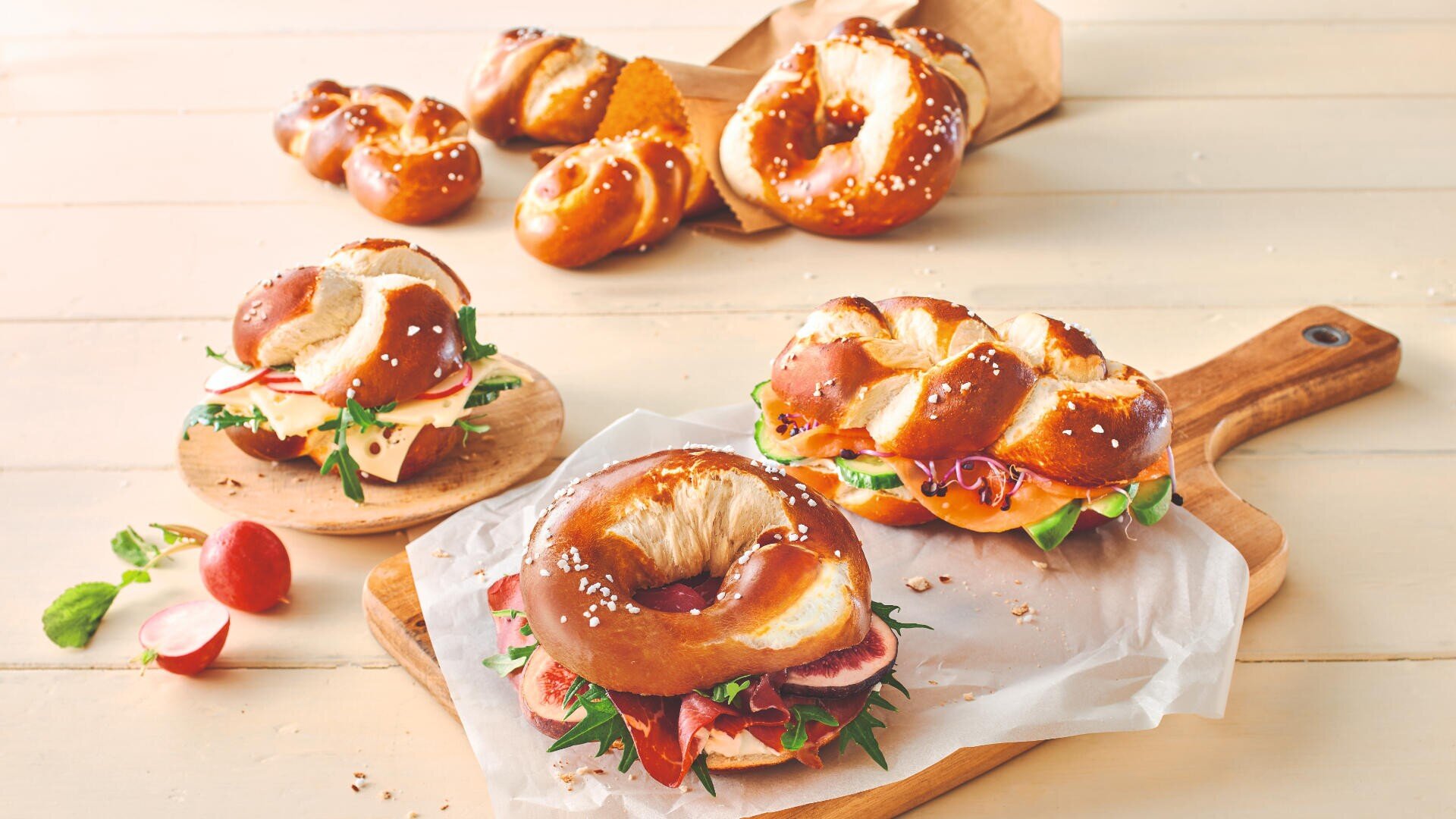 Ein Holztisch mit Brezel-Sandwiches, gefüllt mit Salat, Käse und Aufschnitt, auf einem Schneidebrett und Backpapier. Auch ganze Brezeln und Radieschen liegen verstreut herum.