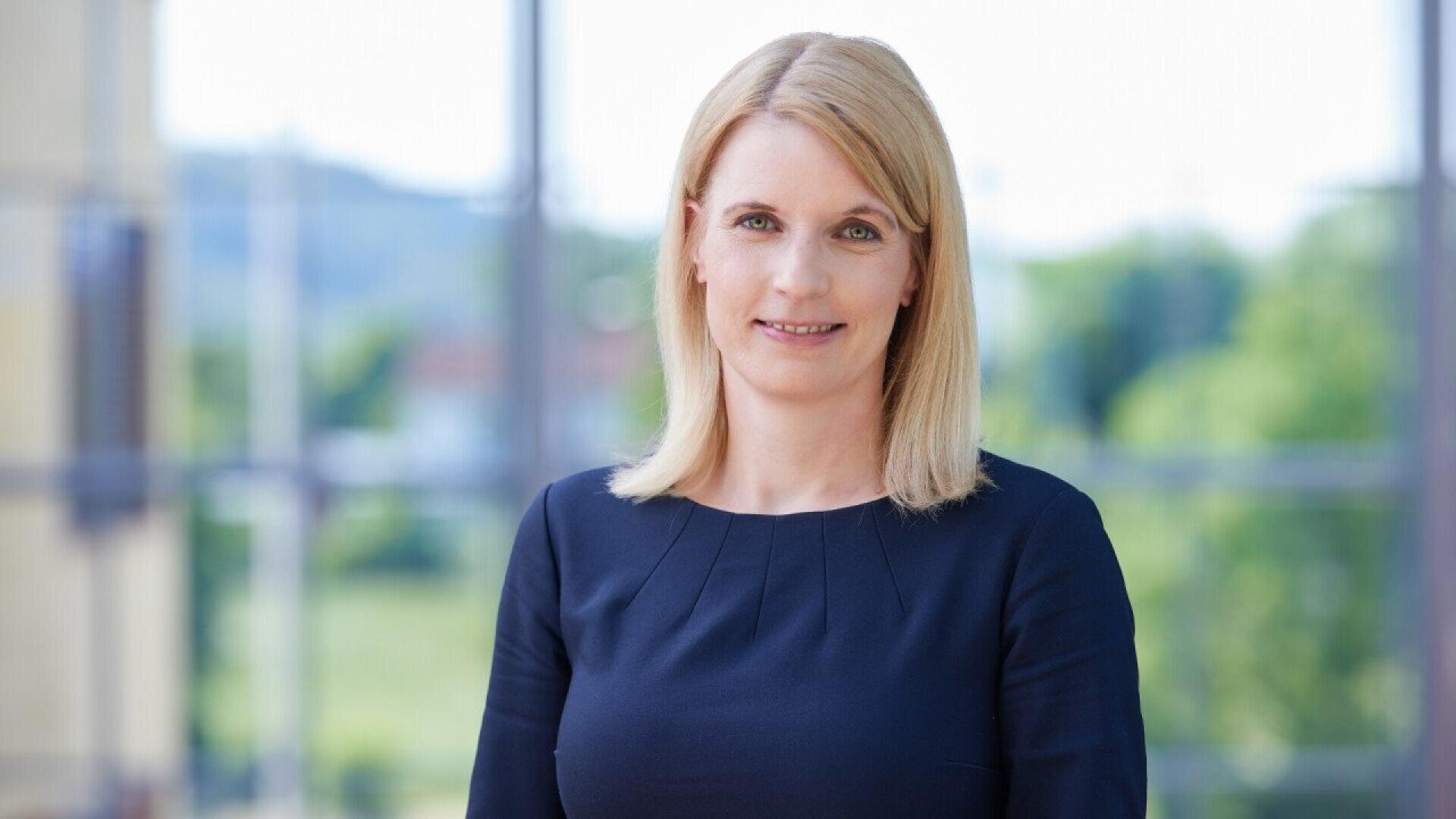 A woman with straight blonde hair and light skin is standing indoors in front of large windows. She is wearing a dark blue top and smiling slightly, with a blurred green outdoor scene visible in the background.