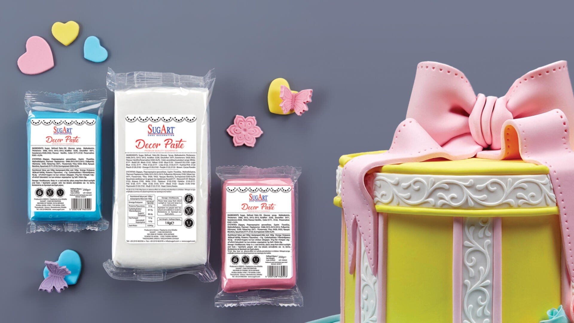 Three packs of Sugar Paste in blue, white, and pink are displayed next to a decorated cake with a large pink bow and yellow sides. Colorful heart and butterfly decorations are scattered on the gray surface.