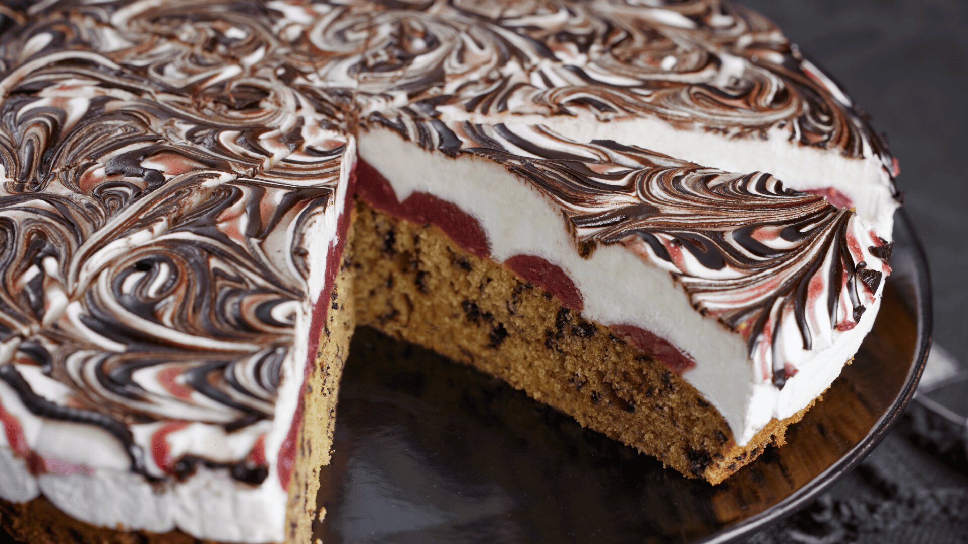 Ein runder Kuchen, bei dem eine Scheibe entfernt wurde, mit Schichten aus Schokoladenbiskuit, roter Fruchtfüllung und dicker weißer Sahne, die mit marmorierter Schokolade und roten Strudeln verziert ist.