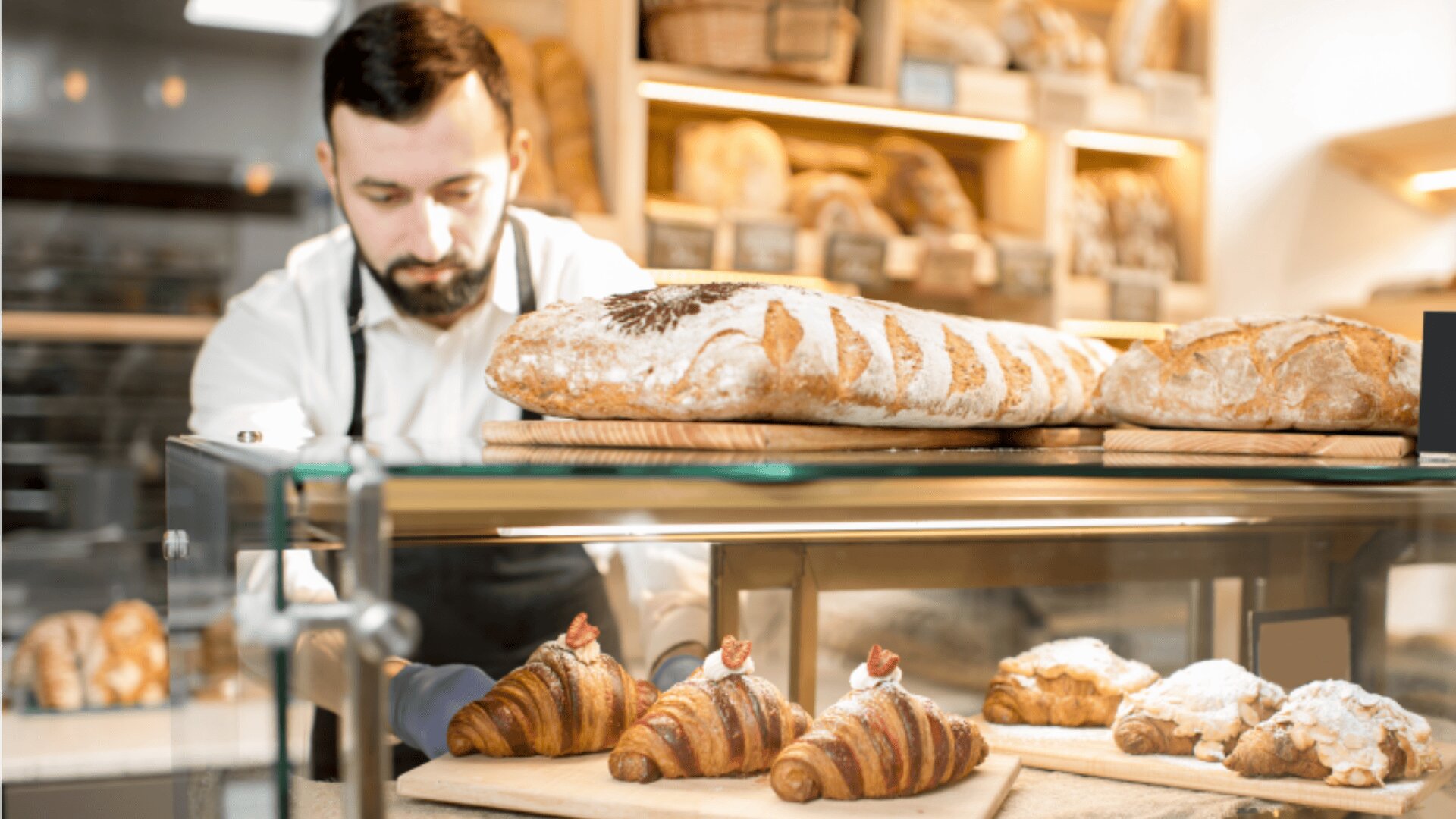 A baker arranges fresh croissants and pastries in a glass display case at a bakery, with loaves of bread on wooden shelves behind him.