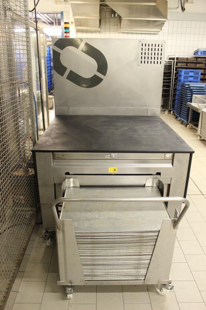 A commercial kitchen setting with a stainless steel oven or warming unit, a pulled-out tray rack in front, and stacked trays visible. Blue racks and tiled walls are in the background.