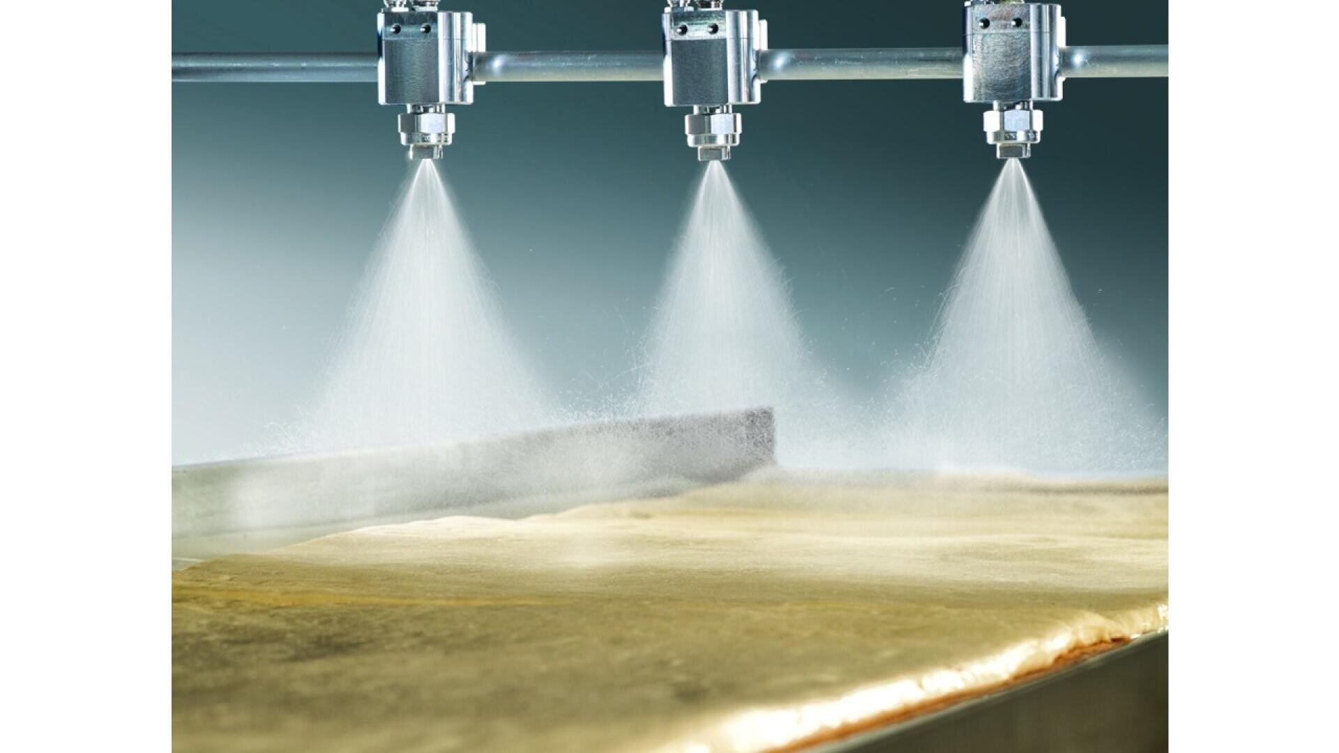 Three metal spray nozzles release a fine mist over a large, flat, yellowish surface, possibly in an industrial or food processing setting. The background is a gradient blue-green color.