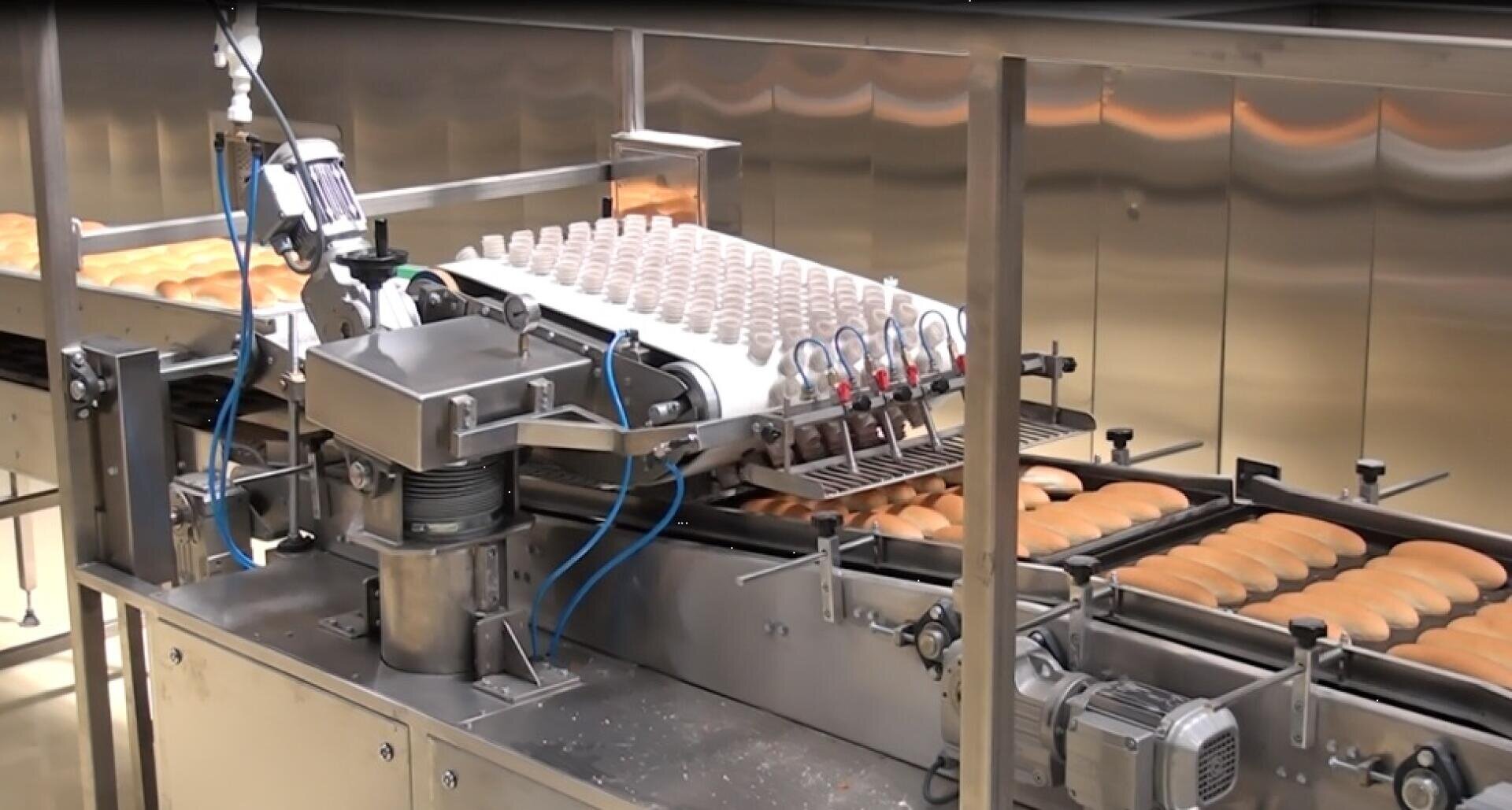 Eine Bäckerei-Produktionslinie mit einer Maschine, die Sahne auf Reihen von Biskuitkuchen aufträgt, die sich auf einem Förderband in einer industriellen Umgebung aus Edelstahl bewegen.