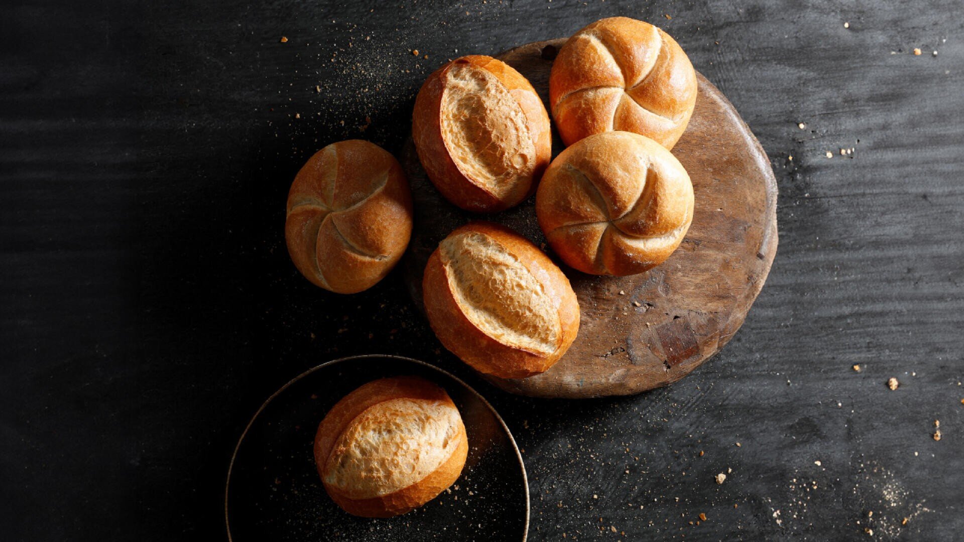 Sechs runde Brötchen, manche mit gewirbelter Oberseite und manche mit einem einzigen Schlitz in der Mitte, liegen auf einem Holzbrett und einer dunklen Oberfläche, rundherum sind Krümel verstreut.